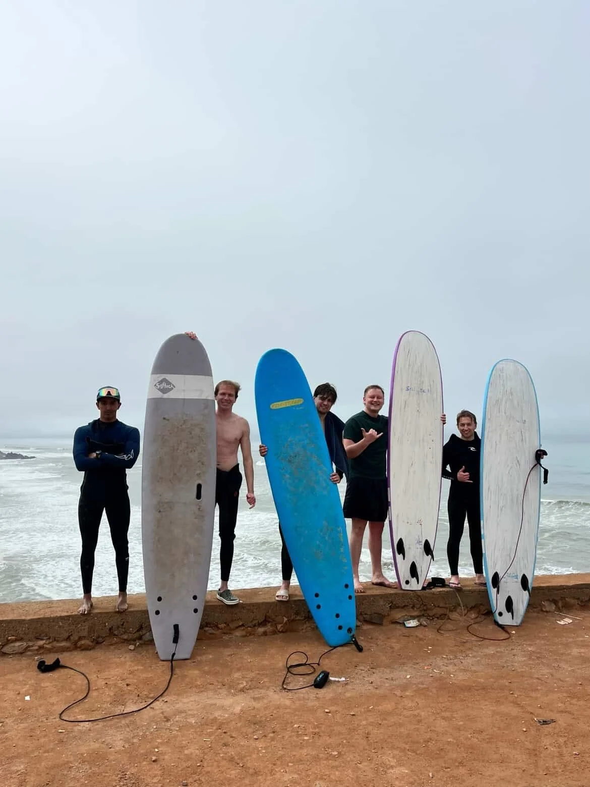 Five men standing on a beach with surfboards, the ocean and cloudy sky in the background. Some men are smiling and posing, with a few making hand gestures. The surfboards are different colors and sizes, and there are surfboard leashes on the ground.