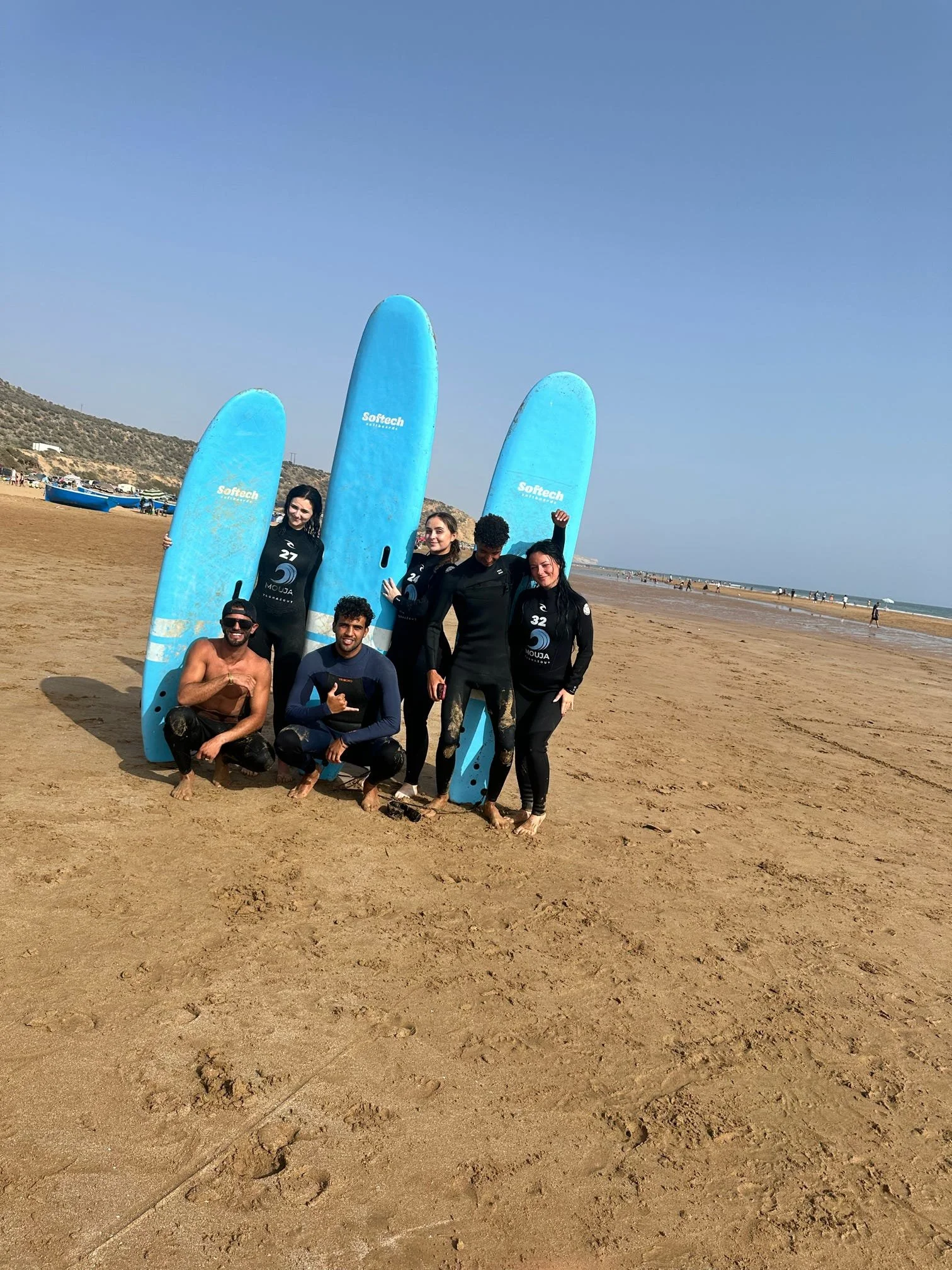 A group of six people standing and kneeling on a beach, holding surfboards and smiling, with the ocean and distant hills in the background on a sunny day.