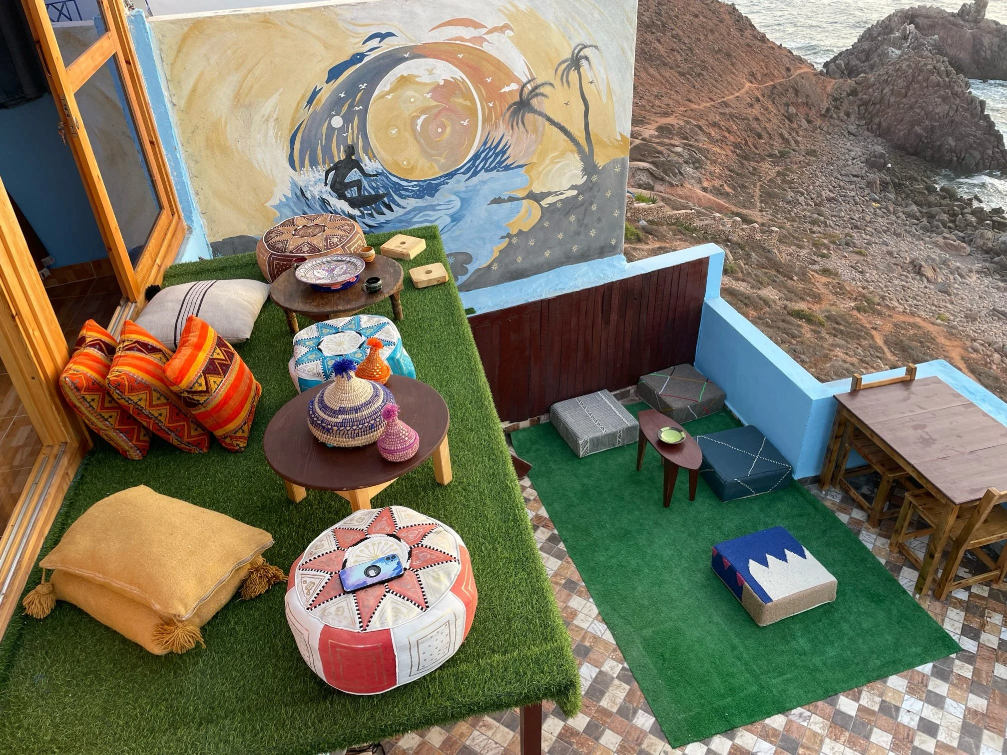 An outdoor terrace overlooking rocky cliffs and the ocean. The terrace has colorful cushions, a small round wooden table with decorative items, a bean bag, and a mural of a surfer riding a wave with palm trees.