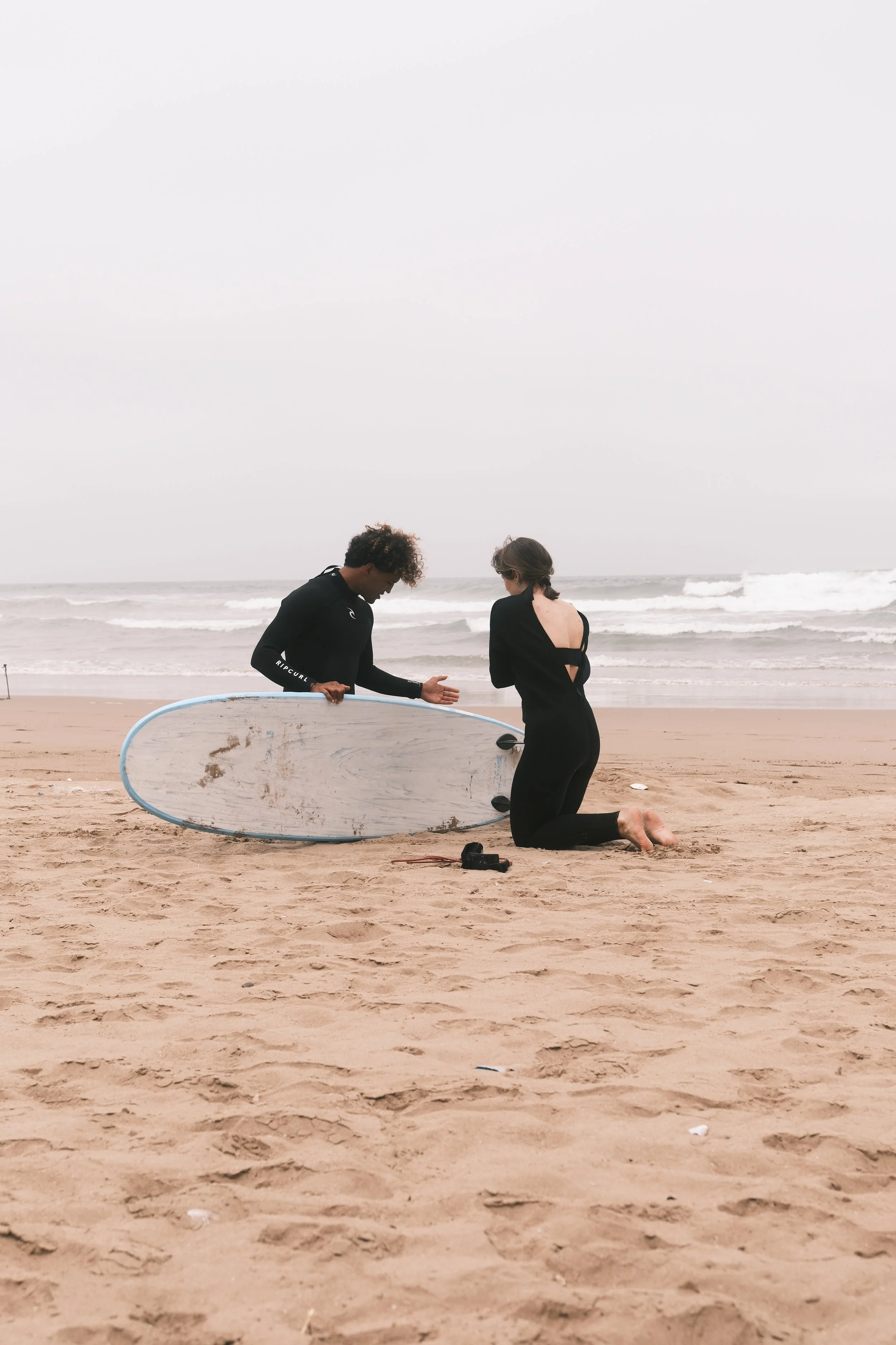 A man kneeling on the sand in front of a woman at the beach, holding out his hand. The woman is kneeling, wearing a black swimsuit with an open back. There is a surfboard on the ground beside them, and the ocean waves are visible in the background un