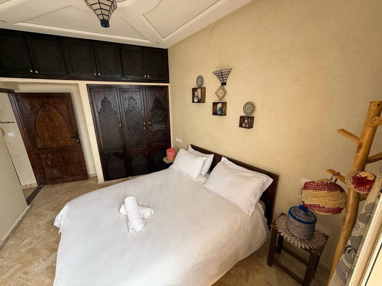 A bedroom with a neatly made bed, white bedding, and pillows. There are towels rolled on the bed. Dark wooden closet and door, decorative wall shelves with ornaments, and a small bedside table with a basket and decorative items. The room has beige wa