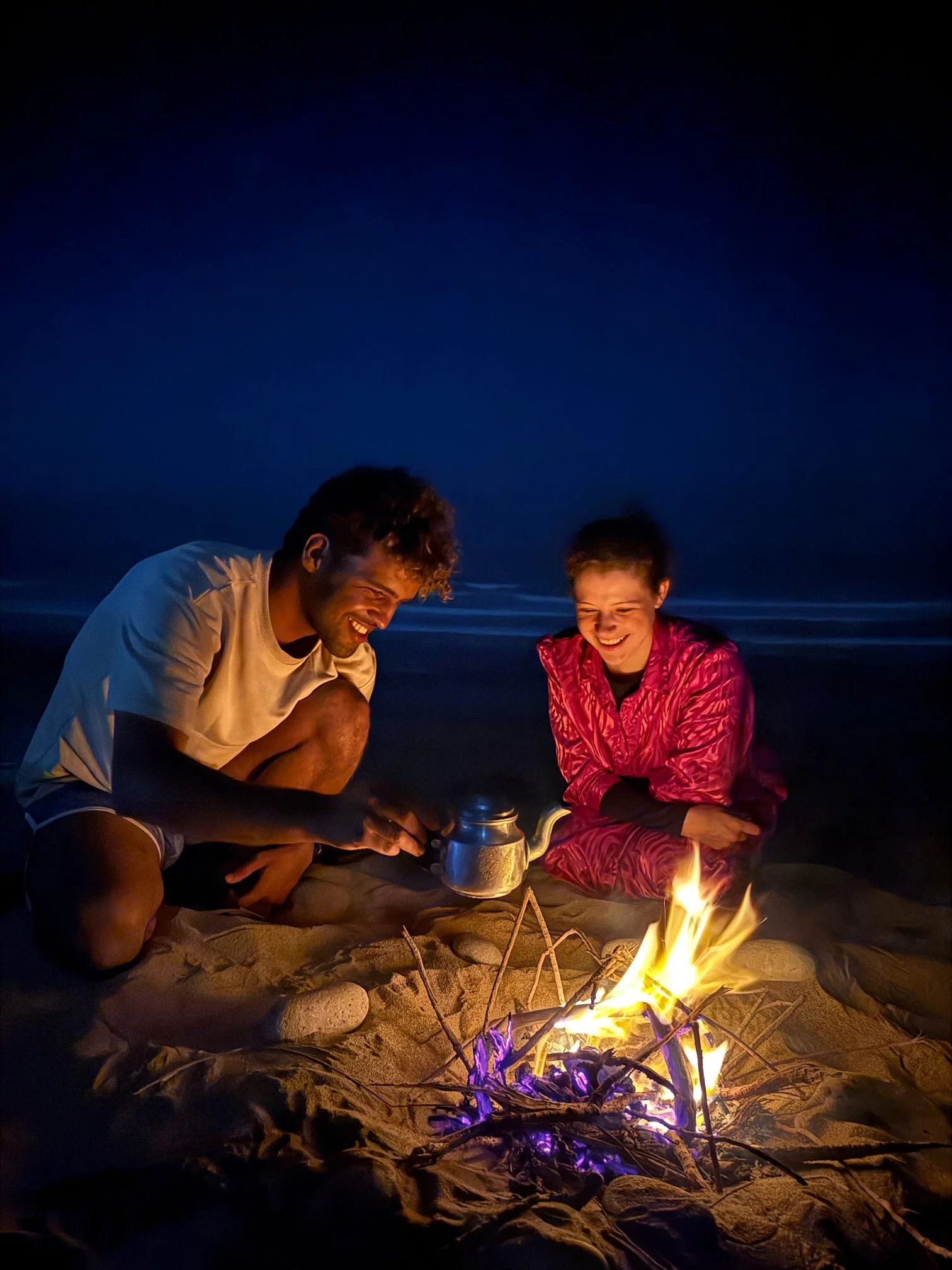 A man and woman sitting on a beach at night around a campfire, smiling and enjoying each other's company.