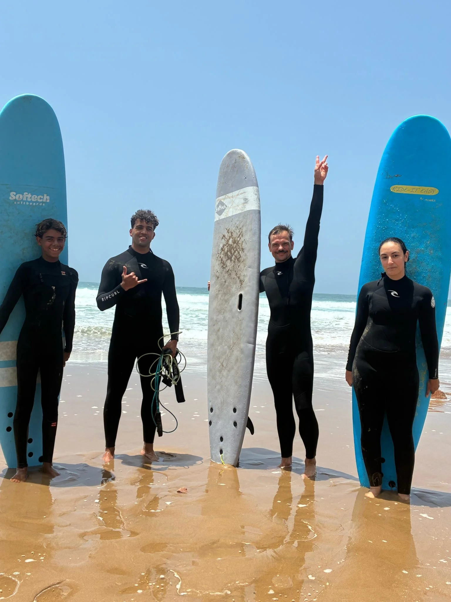 Four people in wetsuits standing on the beach holding surfboards, with one person making a peace sign. They are near the ocean with waves behind them.