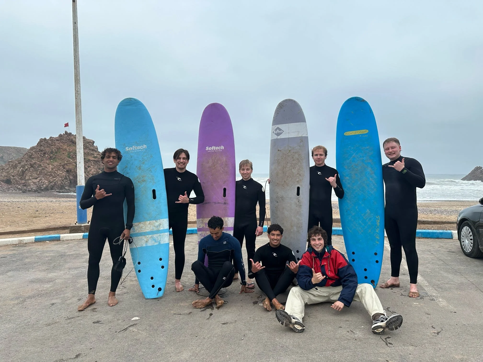 Group of young people at the beach holding surfboards and making shaka signs, with cloudy sky and rocky coast in the background.