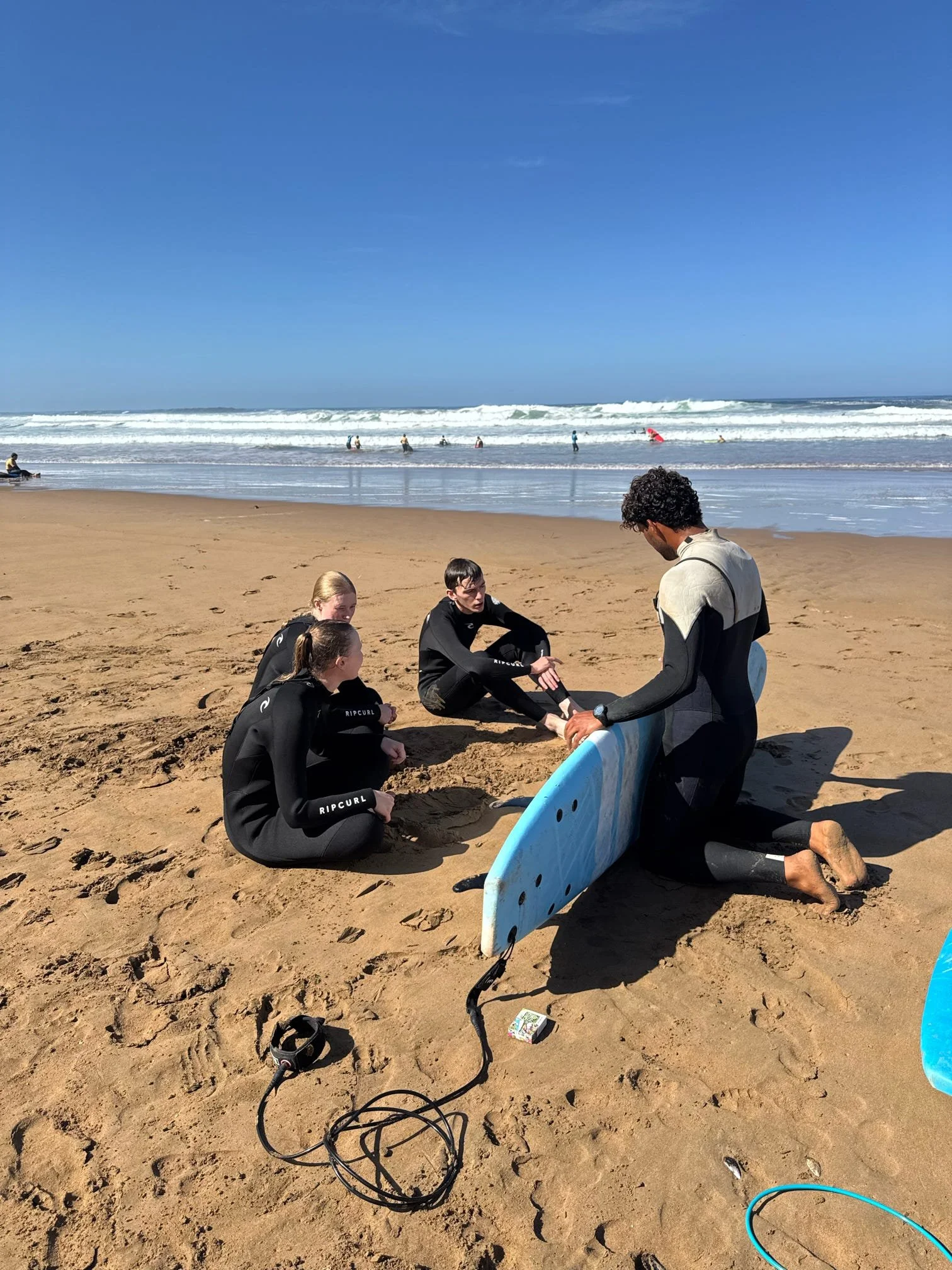 A group of people in wetsuits gathered on a sandy beach, with some sitting and others kneeling, as a surf instructor teaches them how to surf with a surfboard. The ocean waves roll in the background under a clear blue sky.