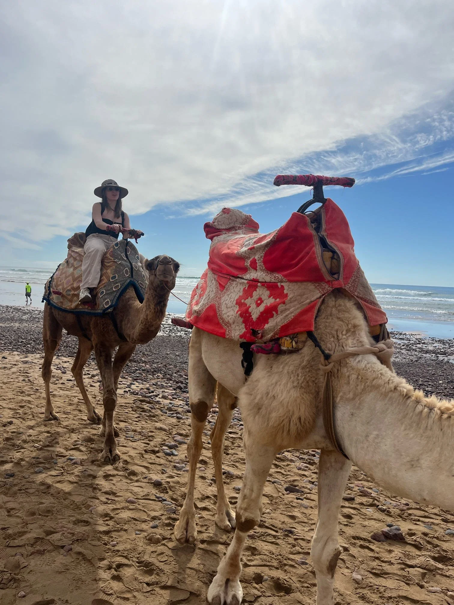 A woman riding a camel on a sandy beach with the ocean and sky in the background, accompanied by a camel and another person in the distance.