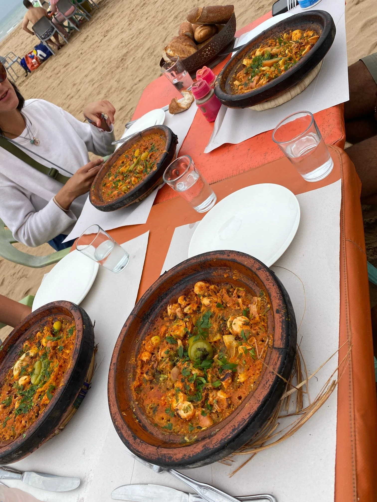 Clams in a tomato-based sauce served in clay pots on a table at a beachside restaurant with bread, water glasses, and salt and pepper shakers.