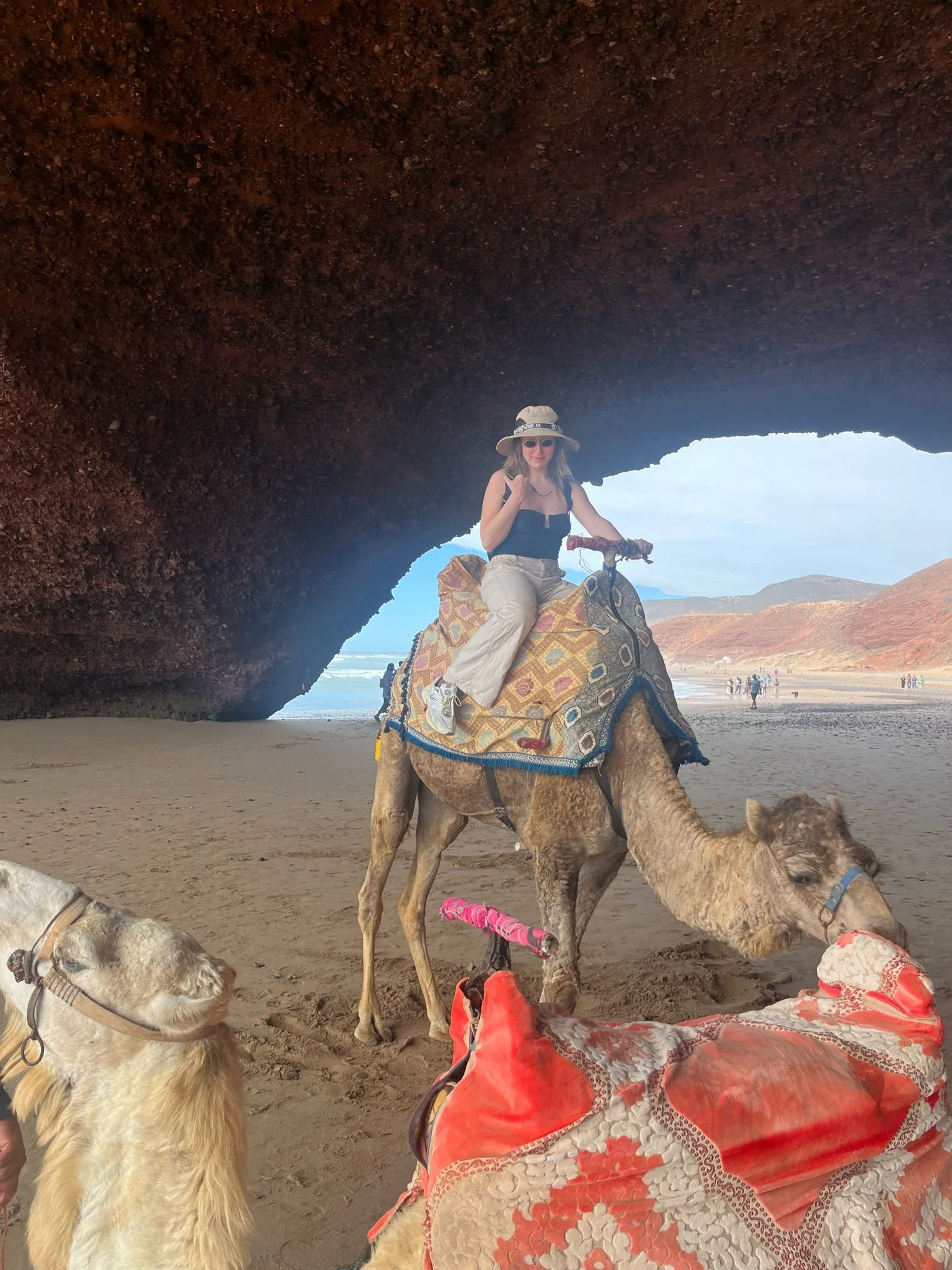 A woman riding a camel inside a large rock formation at a beach, with other camels and people visible in the background.