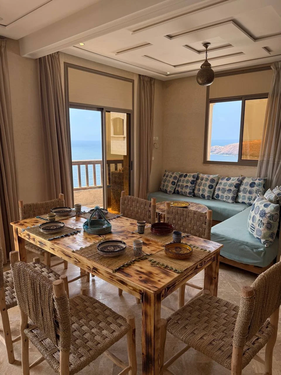A dining room with a wooden table set with decorative plates, cups, and a basket. There are six woven chairs around the table. In the background, there is a sitting area with a sofa adorned with patterned pillows, and large windows showing a view of 