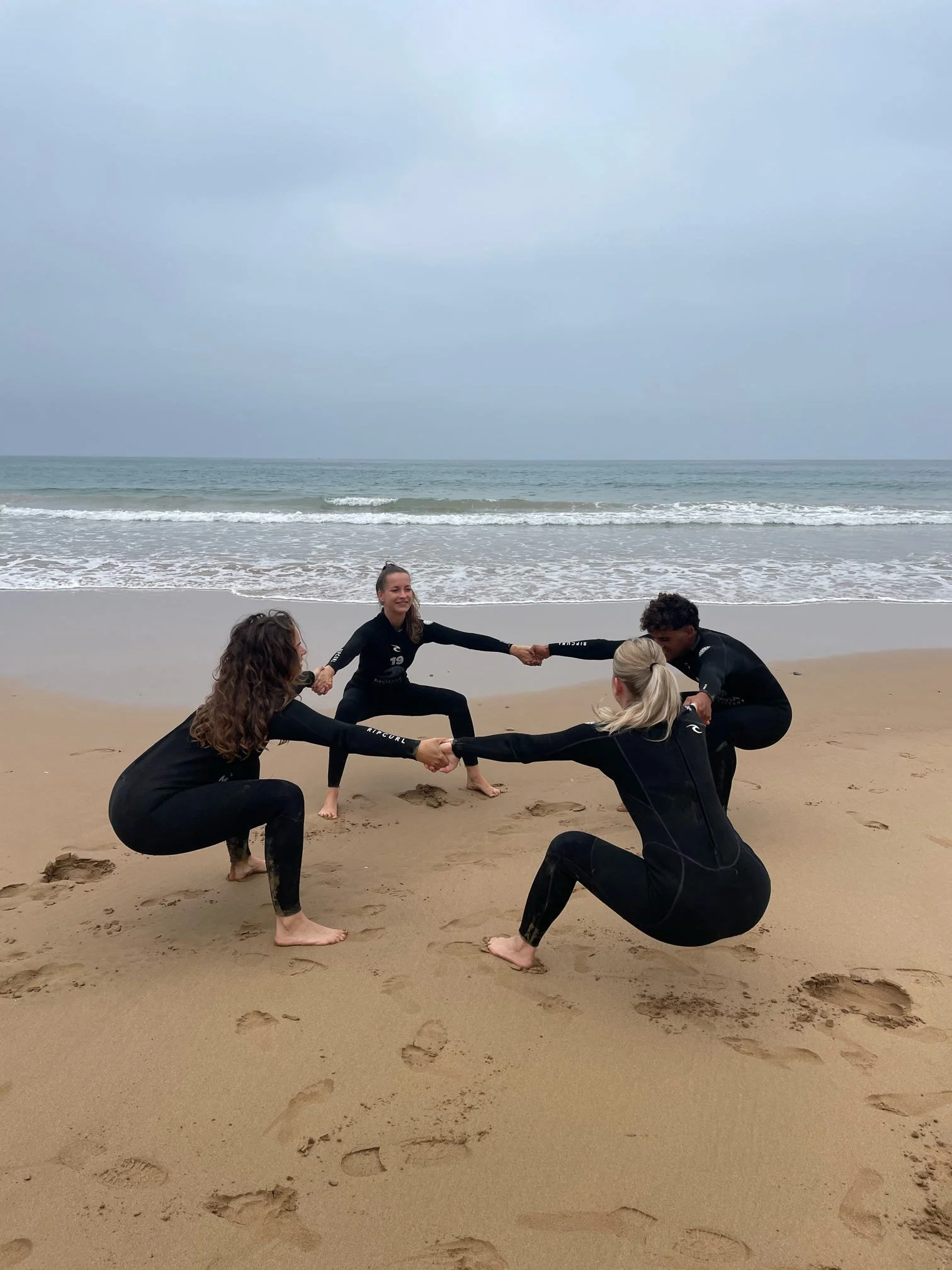 A group of people practicing partner yoga on a beach near the ocean, with cloudy sky and footprints in the sand.