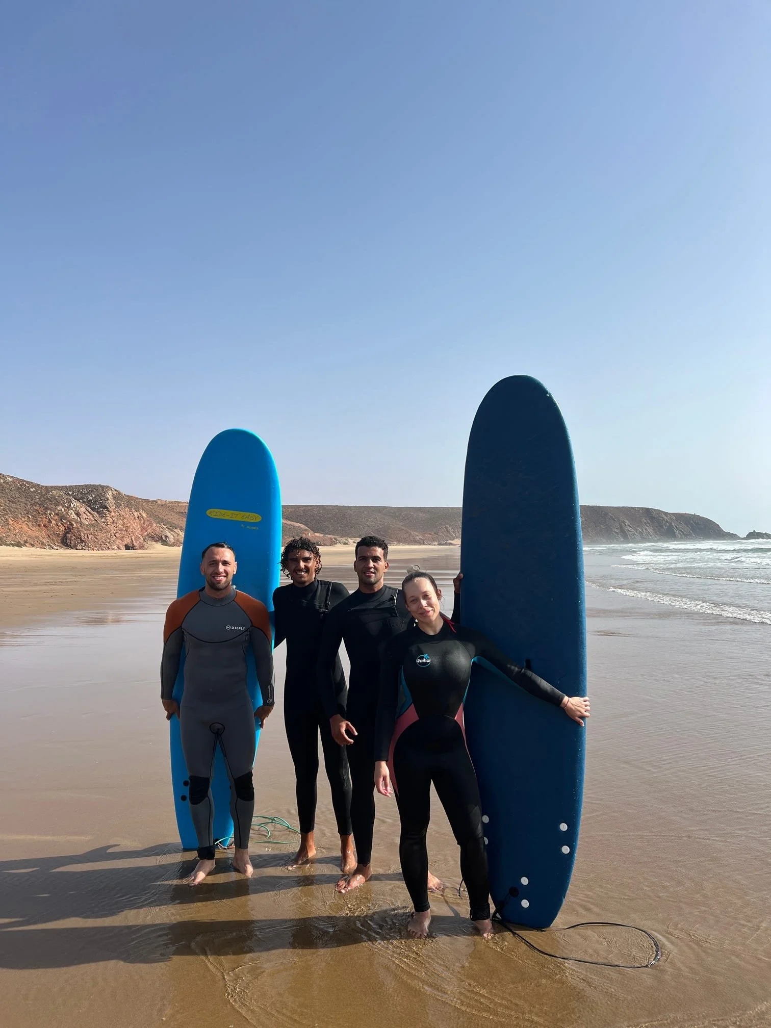 Four surfers standing on a beach with surfboards, wearing wetsuits, at the shoreline with ocean waves and cliffs in the background.