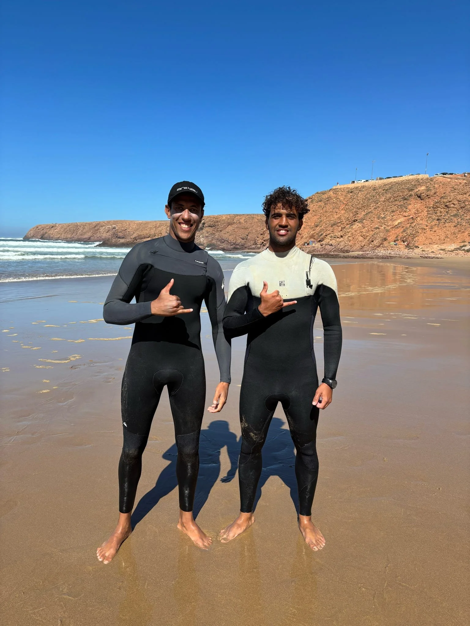 Two men in wetsuits standing barefoot on a sandy beach with water and a rocky hill in the background, smiling and giving thumbs up.