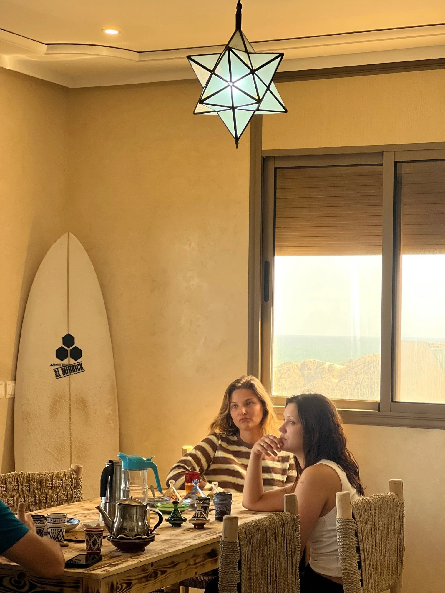 Two young women sitting at a wooden dining table in a cozy room, with large window showing scenic outdoor view, and a geometric hanging lamp above.