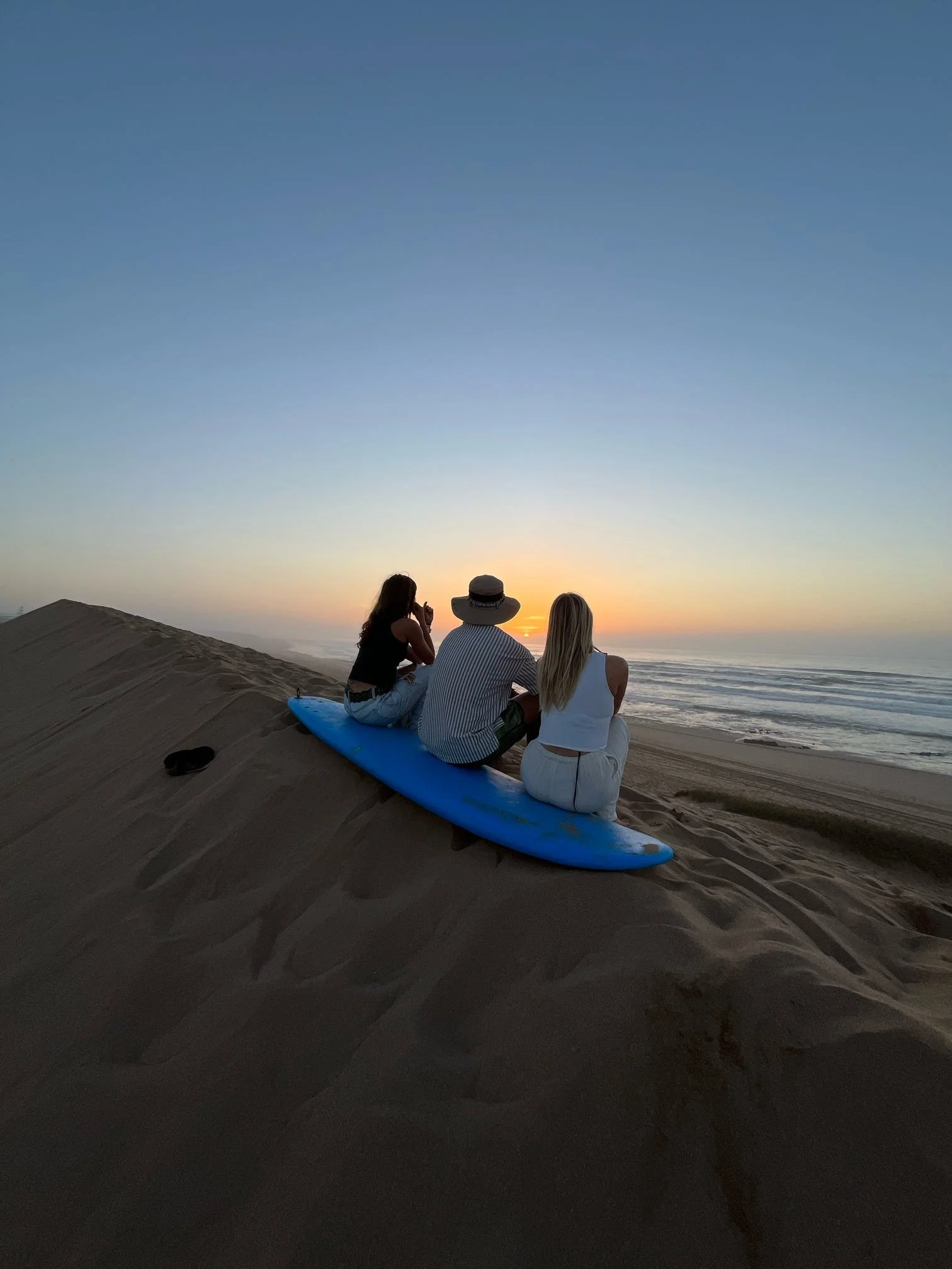 People sitting on the sand at a beach during sunset, with a surfboard nearby, and the ocean in the background.