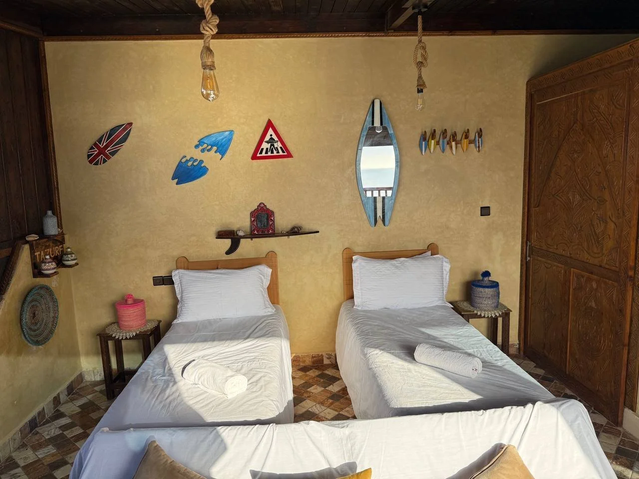 A bedroom with three single beds, each covered with white sheets and pillows, with towels rolled at the foot of each bed. The wall above the beds has decorations including surfboards, a British flag design, a water splash sign, a traffic sign, a mirr