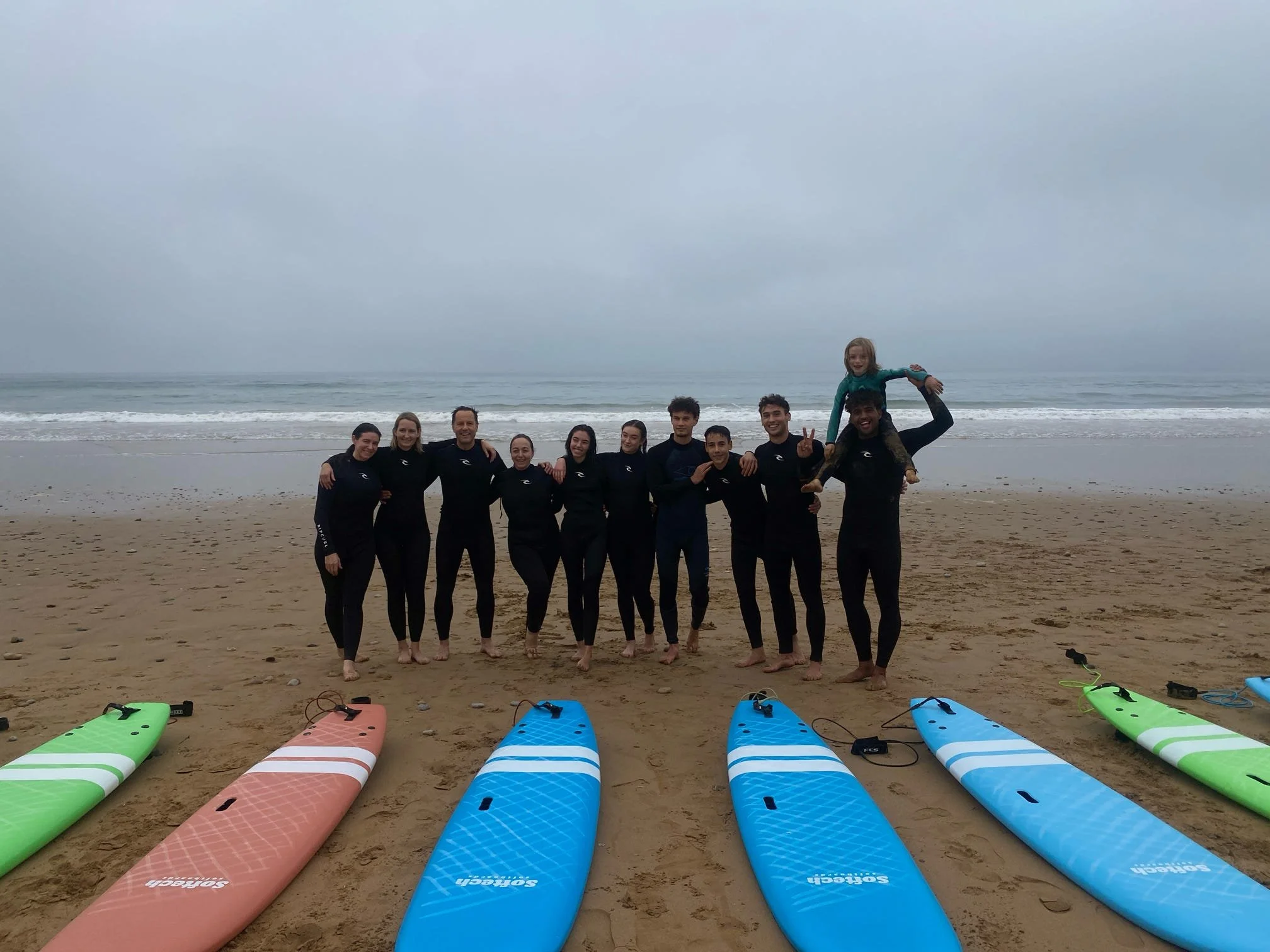 Group of people standing on a sandy beach with surfboards laid out in front of them, overcast sky, ocean in the background.