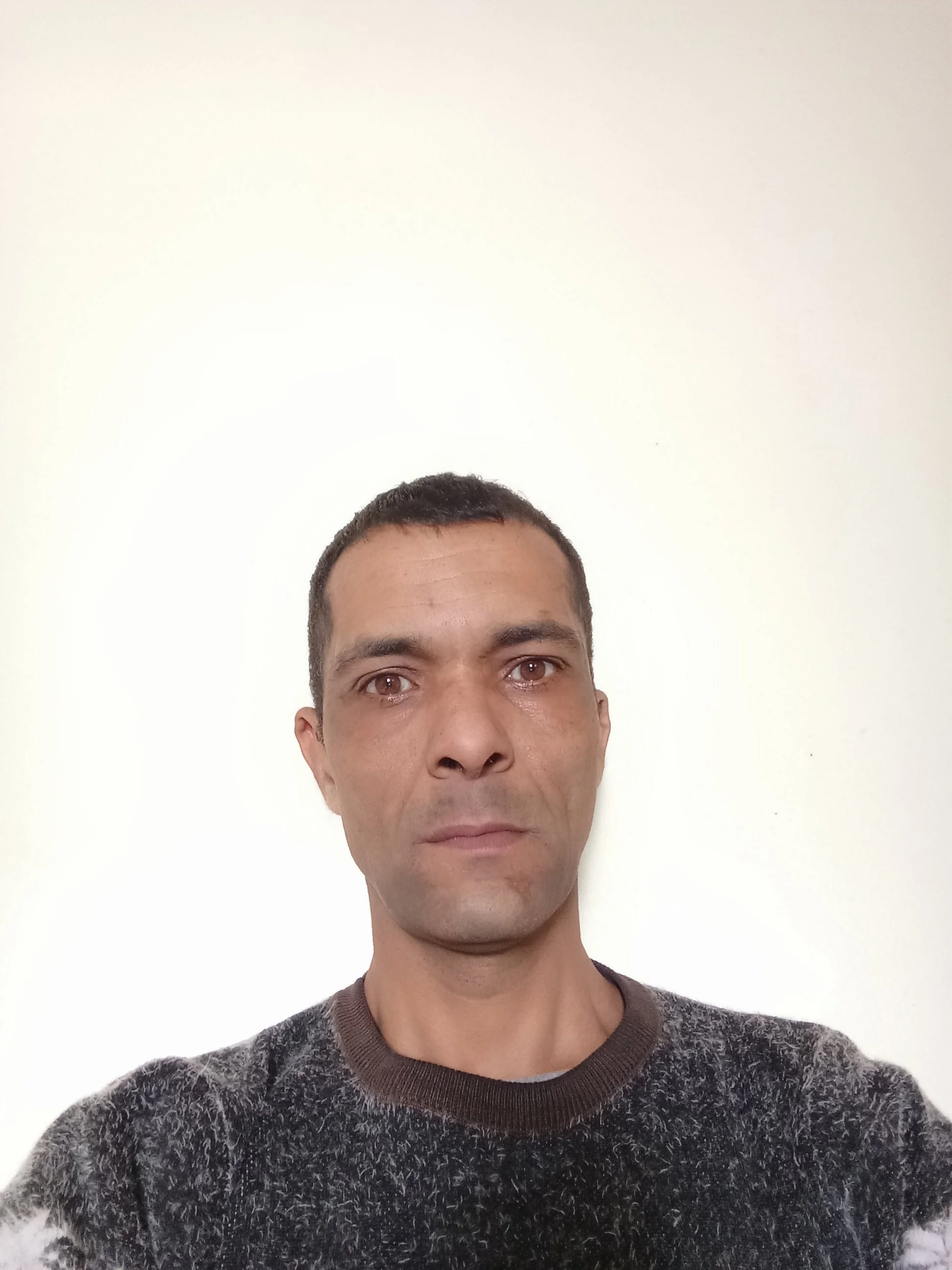 Close-up of a man with short dark hair and brown eyes wearing a patterned gray sweater, standing against a plain white wall.