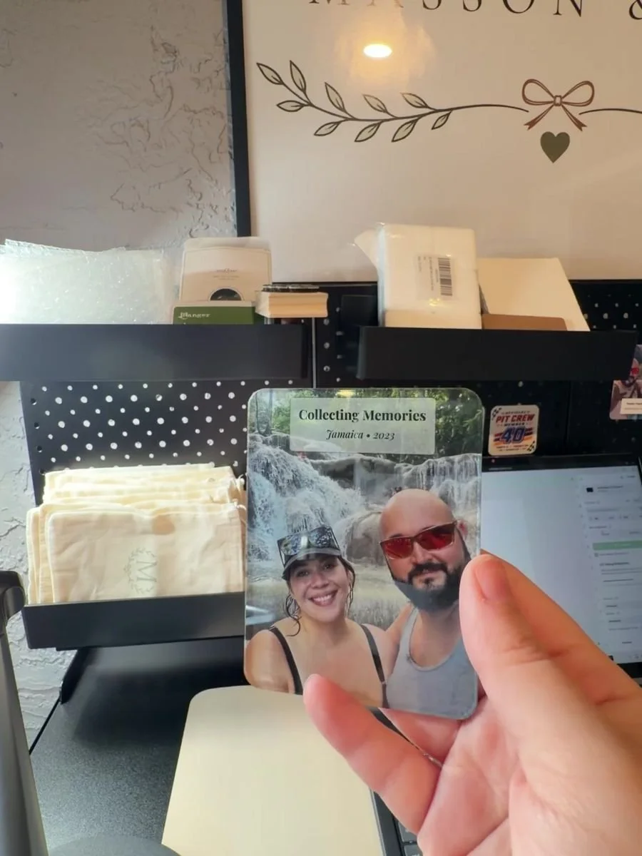 A person is holding a small photo frame with a picture of a smiling couple in front of a waterfall. The background shows a reception area with various pamphlets, documents, and a wall decoration that says 'Collecting Memories Jamaica 2023'.