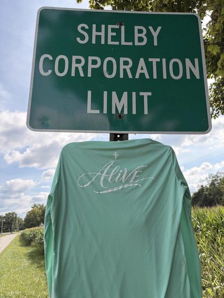 A green road sign reading 'Shelby Corporation Limit' with a cloth draped over the post displaying the word 'Alive' with a cross above it, outdoors on a sunny day.