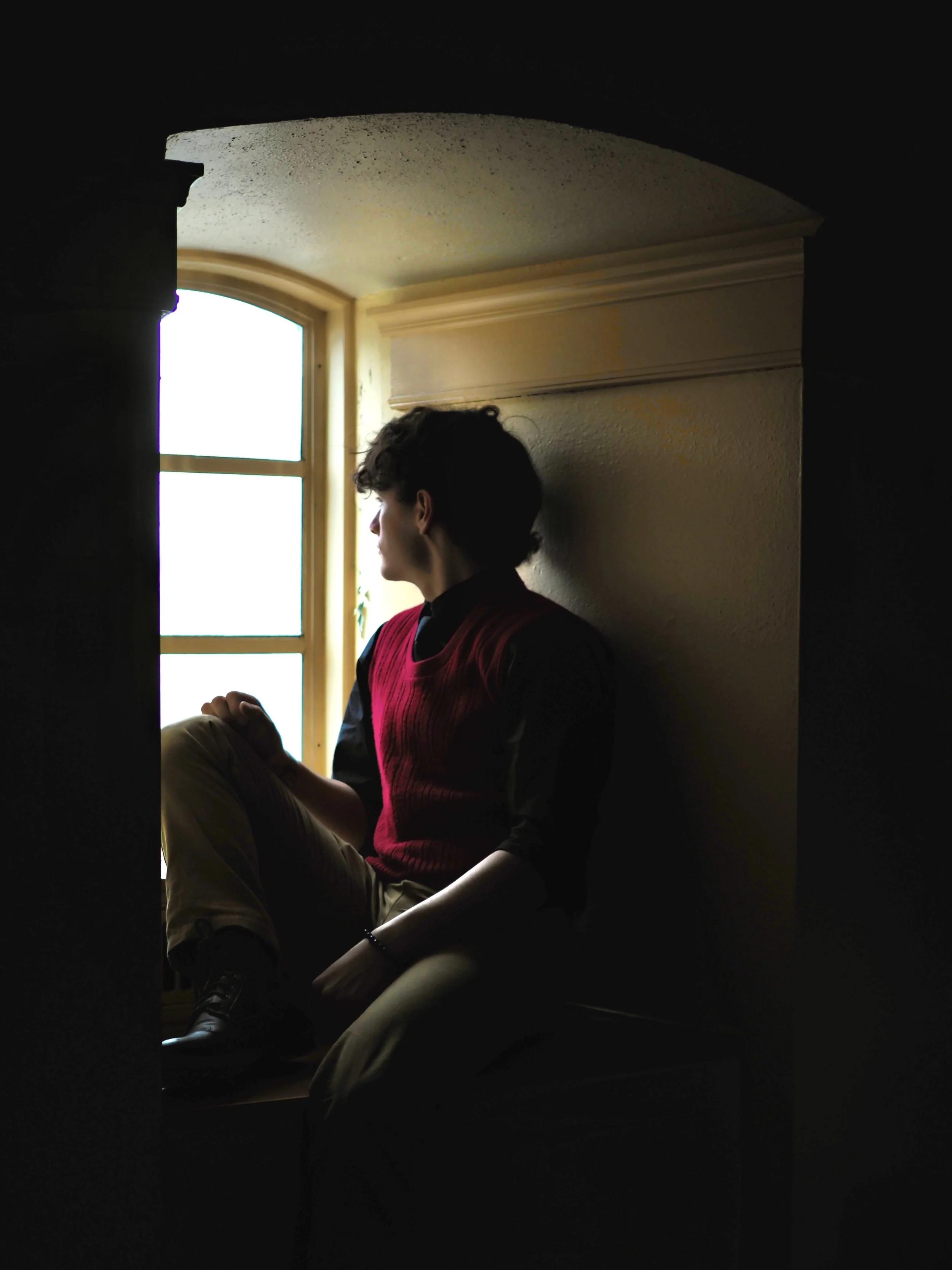 A young man sitting by a window, looking outside, partially silhouetted against the bright daylight. He wears a red vest over a dark shirt and beige pants, with dark hair.