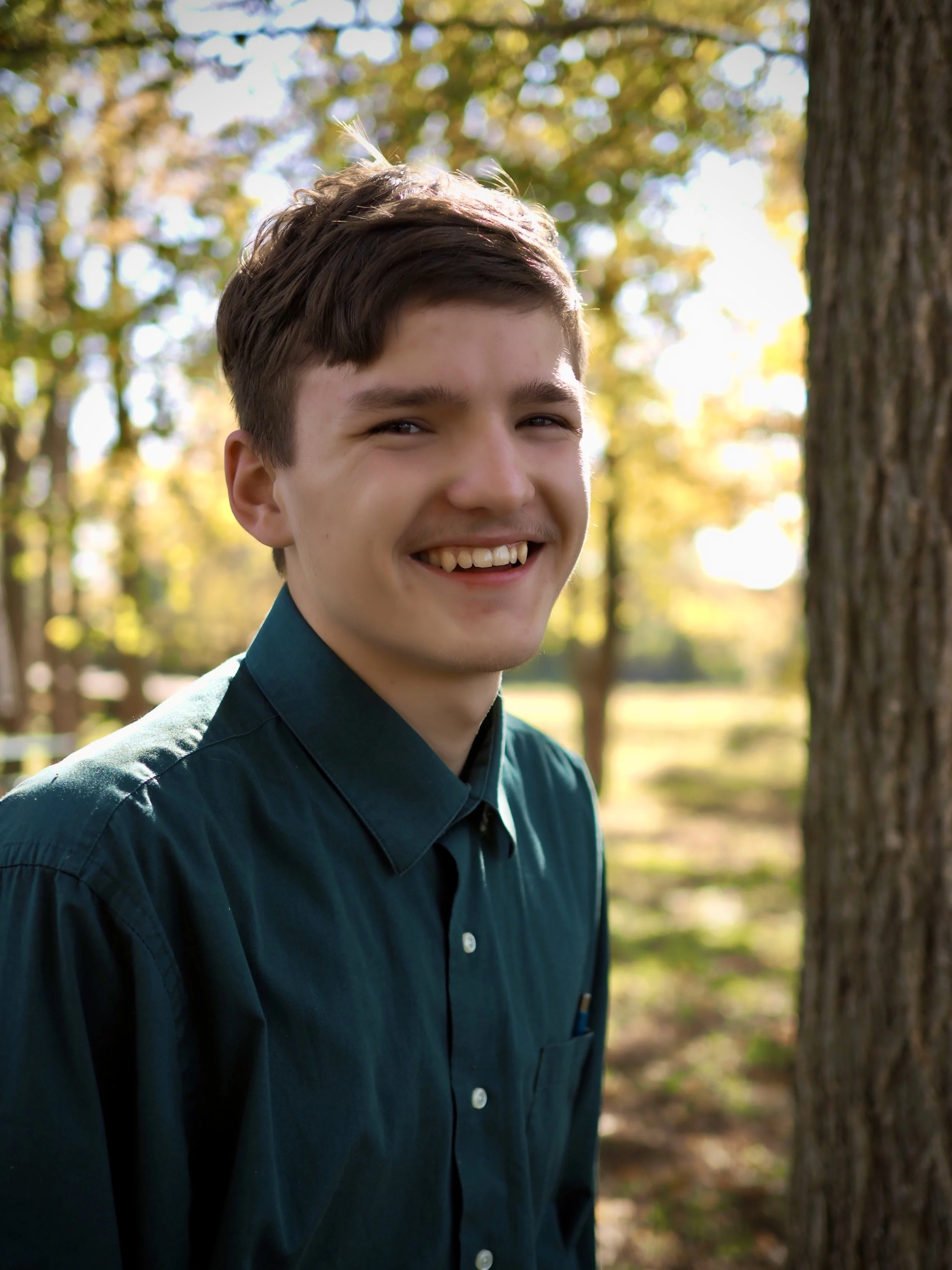 A young man with brown hair smiling outdoors in front of trees with autumn-colored leaves.