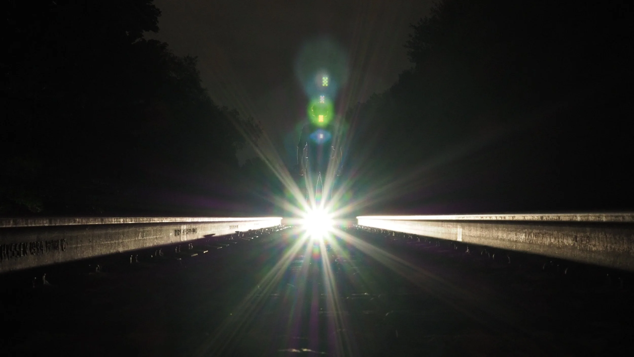 Bright headlight glare on a dark road, creating a starburst effect with a person walking in the distance, flanked by trees.