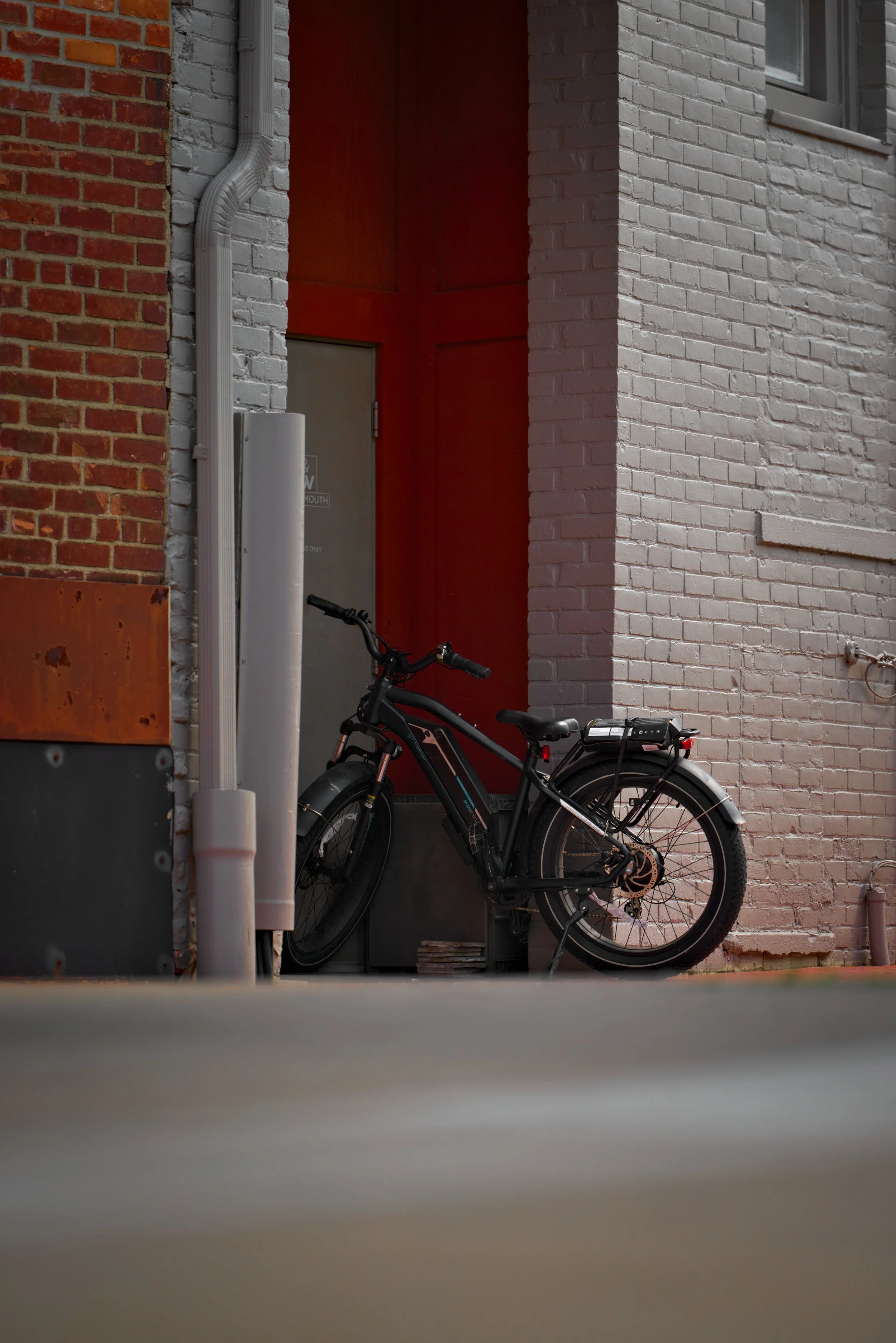 A black electric bicycle parked against a gray door in an alleyway with brick walls, one red and one white, and a white drip pipe attached to the wall.