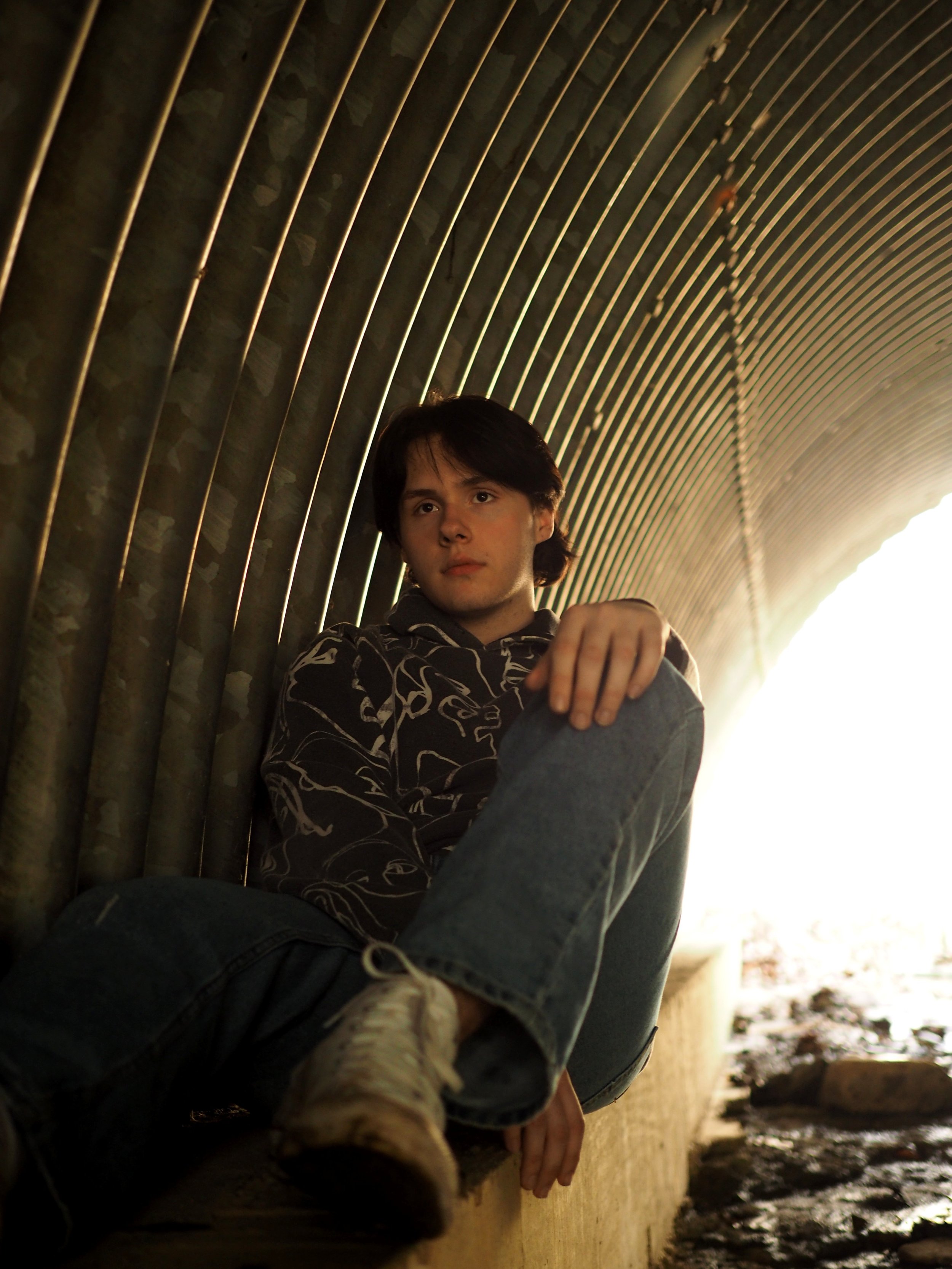 Young man with dark hair sitting against a curved metal wall with vertical bars.