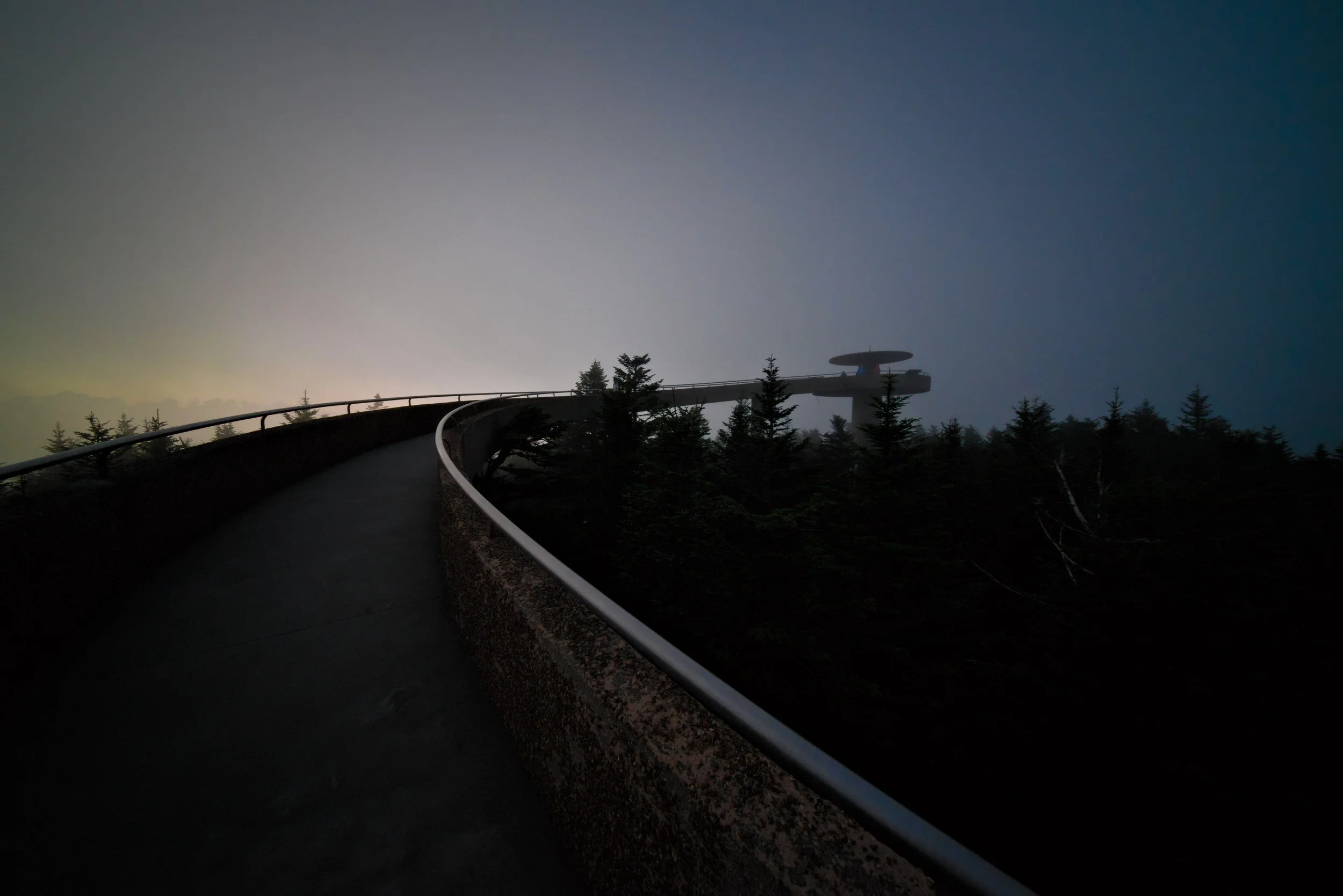 A winding road through a forested area with trees extending into the distance, under a hazy sky with the sun setting or rising.