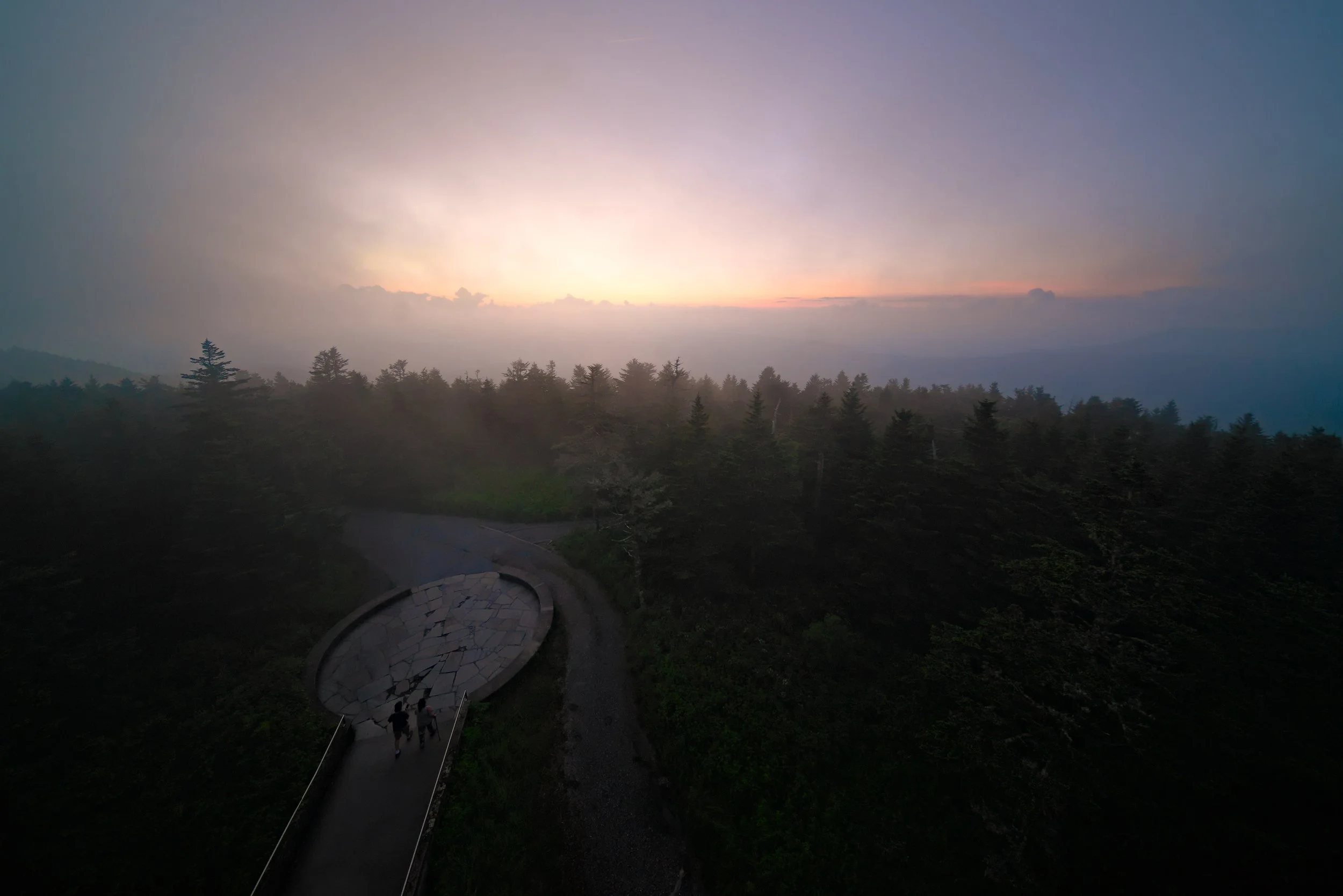 A scenic view of a sunset over a forest, with a winding paved pathway and a couple walking along it.