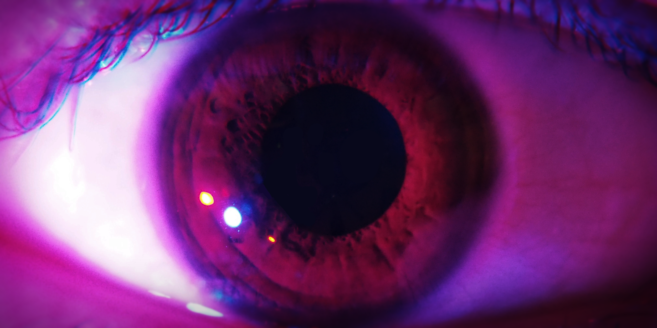 Close-up of a human eye with purple and pink lighting, showing the iris, pupil, and surrounding eye structures.
