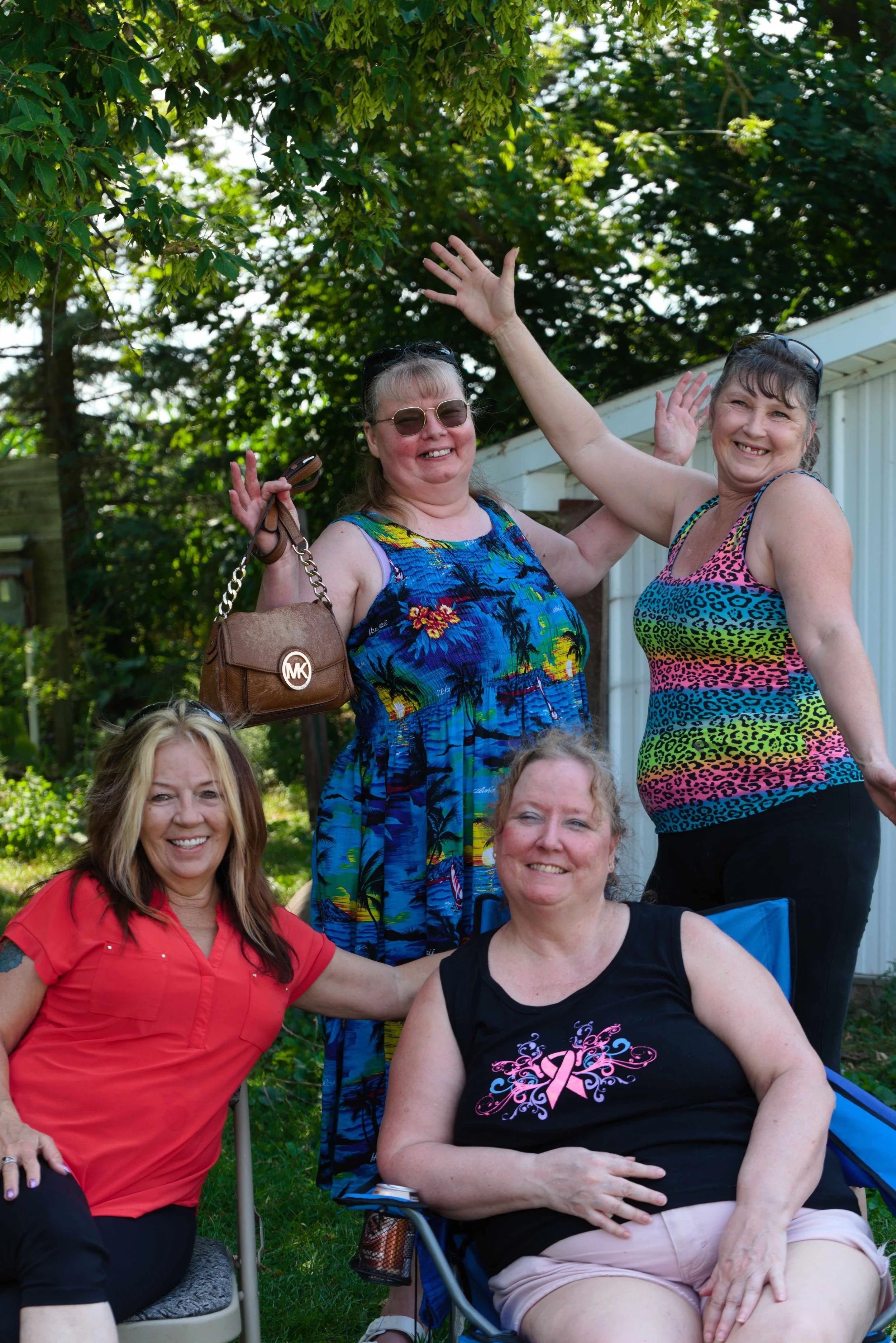 Group of women outdoors, some sitting and some standing, smiling and waving, with trees and a house in the background.