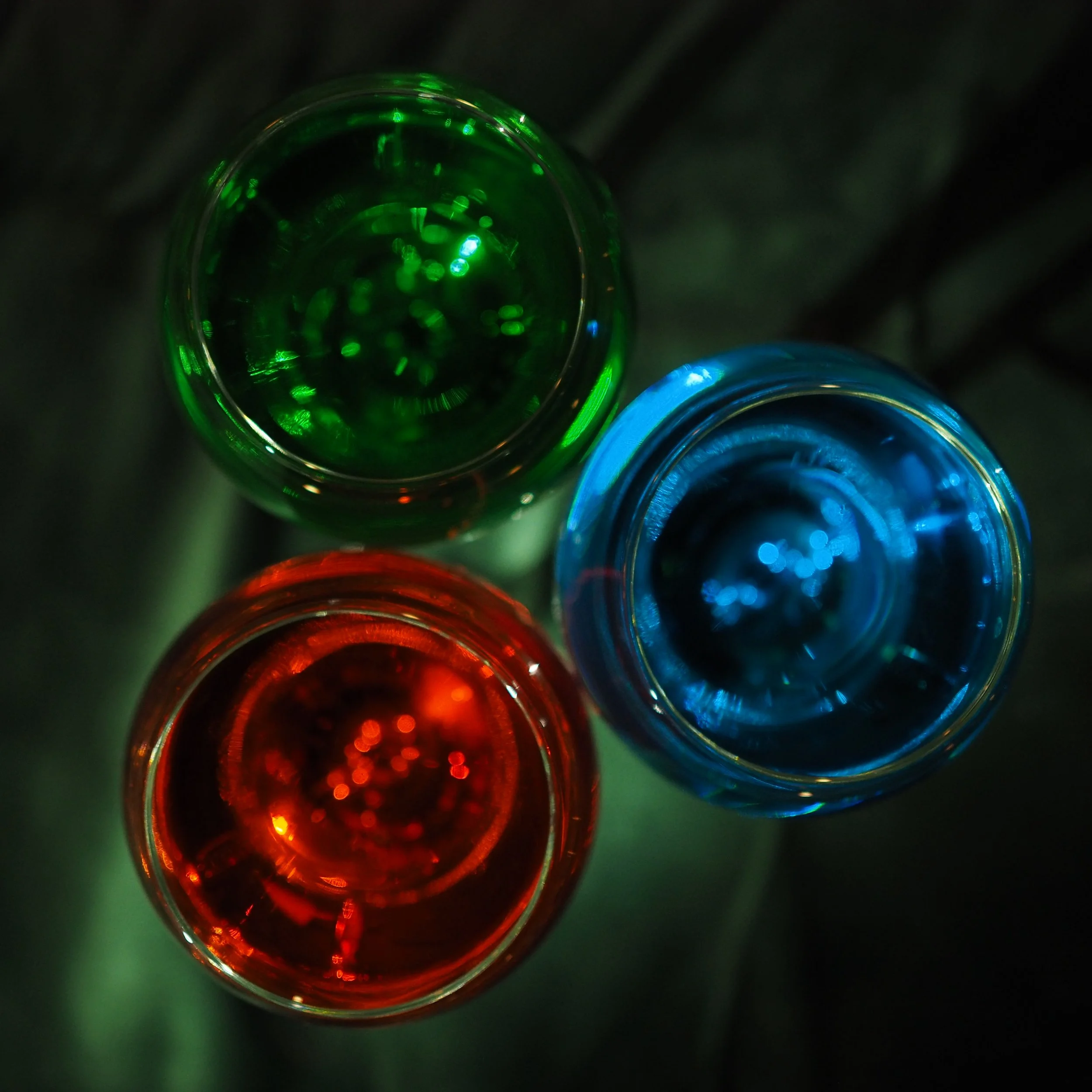 Looking down at the tops of five colored glass bottles arranged in a row, illuminated with different colors of light.