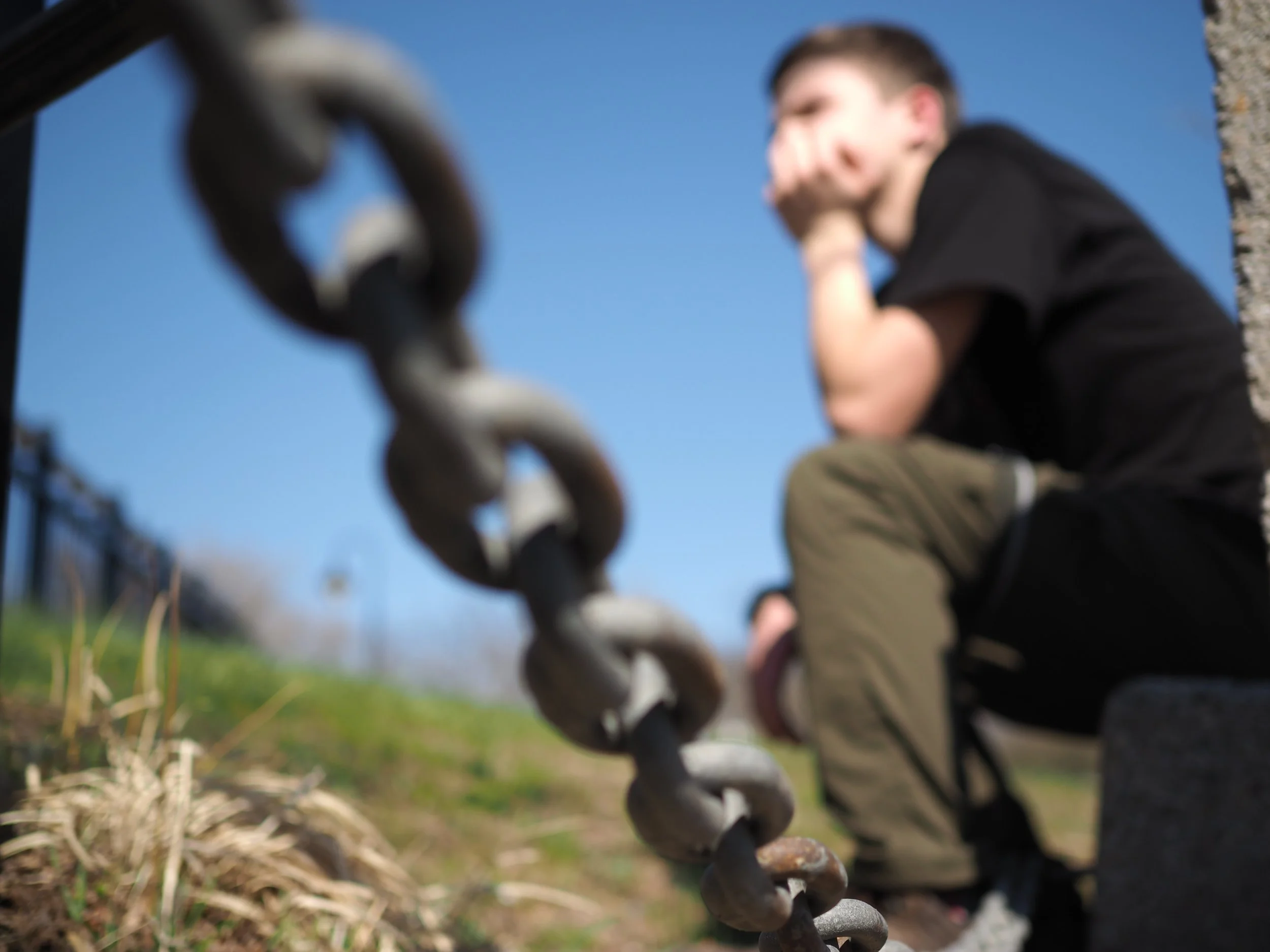 Close-up of a chain link fence with a person in the background, slightly blurred, sitting outdoors against a blue sky.