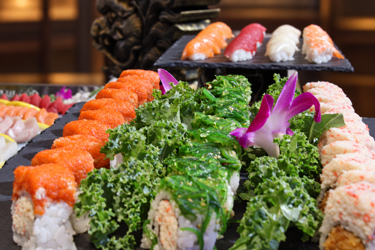 Assorted sushi rolls with green seaweed salad, purple orchids, and fresh greens on a black platter, with additional sushi pieces on elevated black platters in the background.