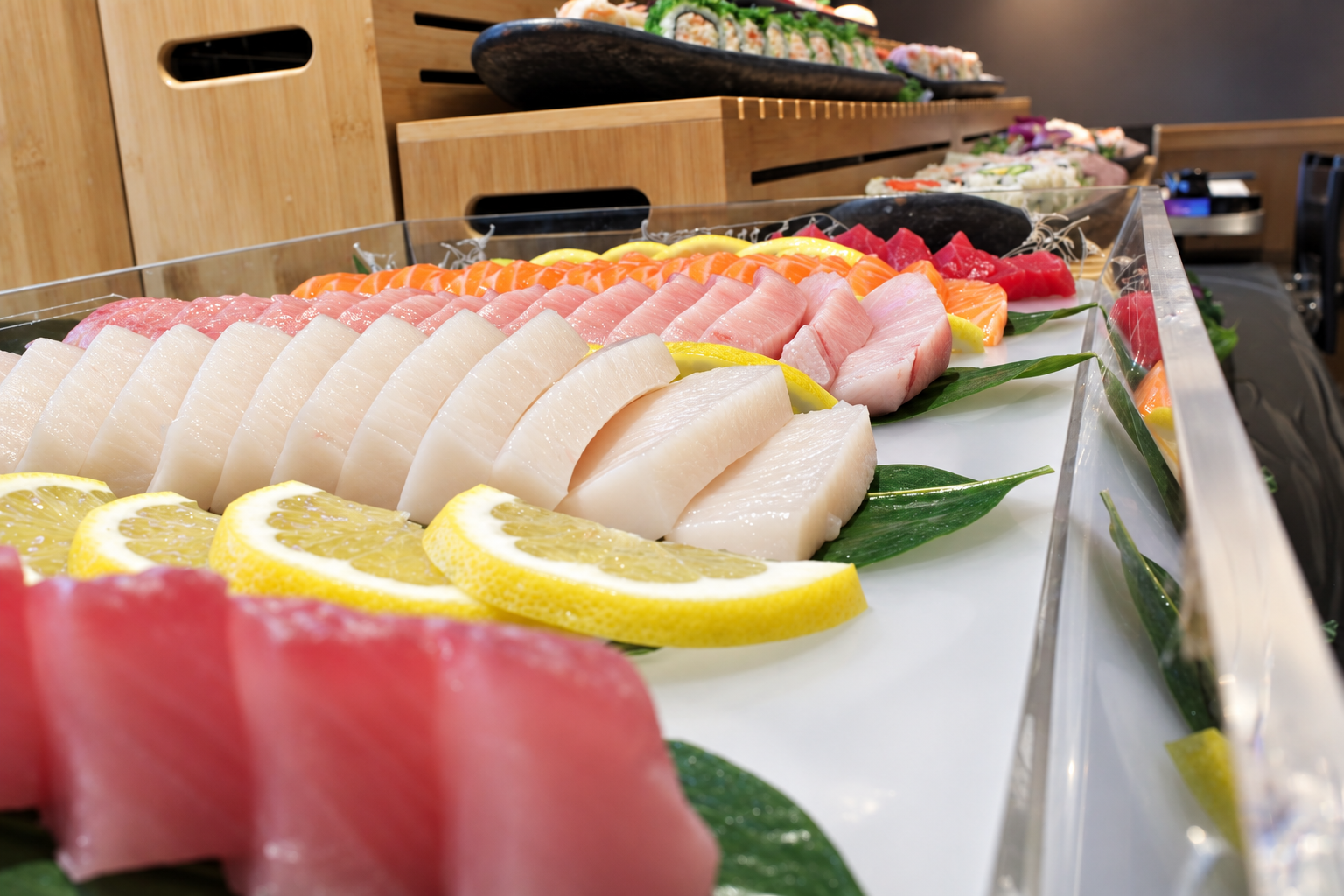 A tray of assorted sashimi including slices of white fish, pink fish, and red tuna, garnished with lemon slices and green leaves, with sushi rolls visible in the background.