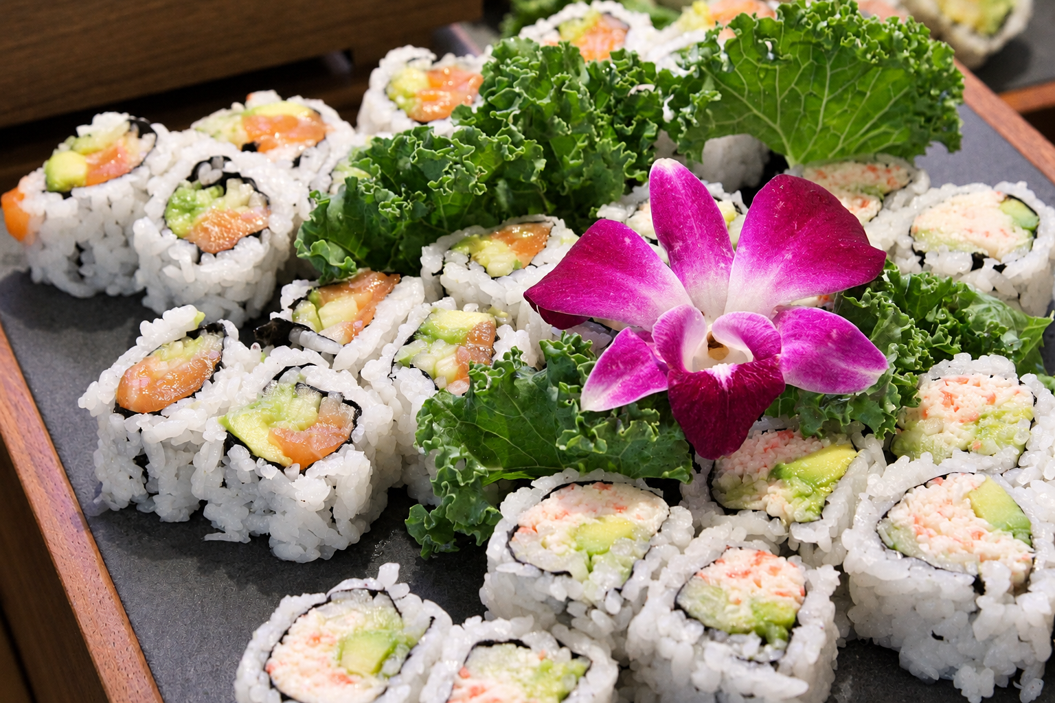 Assorted sushi rolls garnished with kale, an orchid flower, and decorative greens on a black platter.