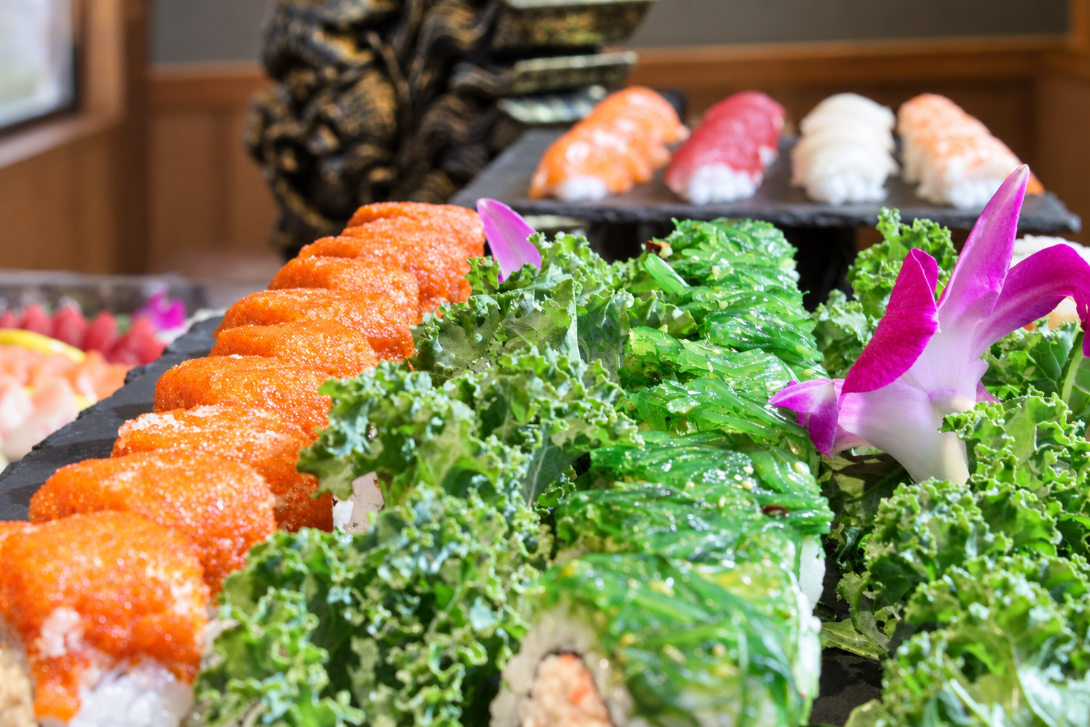 Assorted sushi rolls and sashimi on a platter, decorated with green leafy vegetables and a pink and white orchid flower.