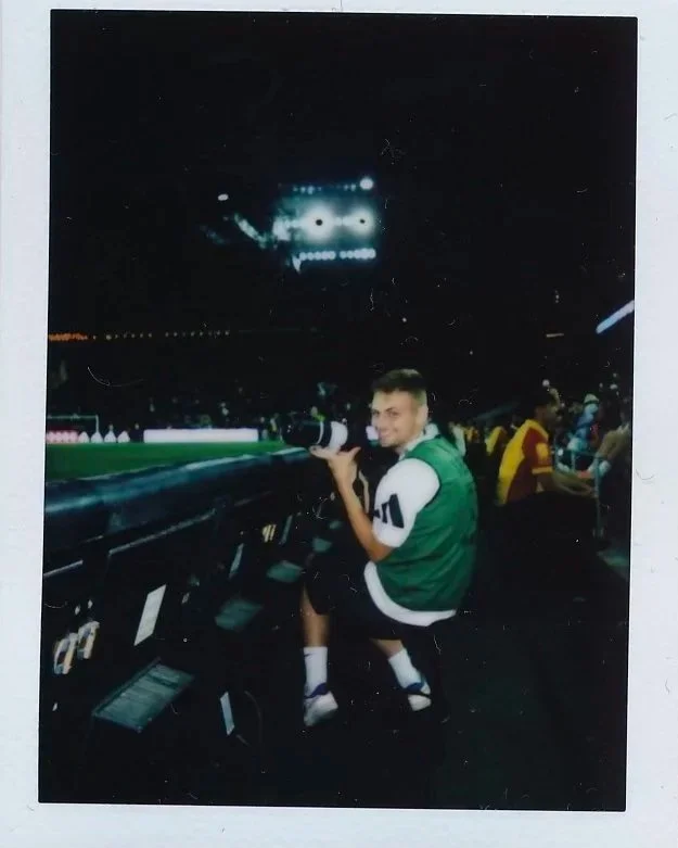 Beau Chevalier at sports stadium sitting on a kickstand with a camera, smiling, wearing a green and white jacket, with other spectators in the background, illuminated scoreboard above.