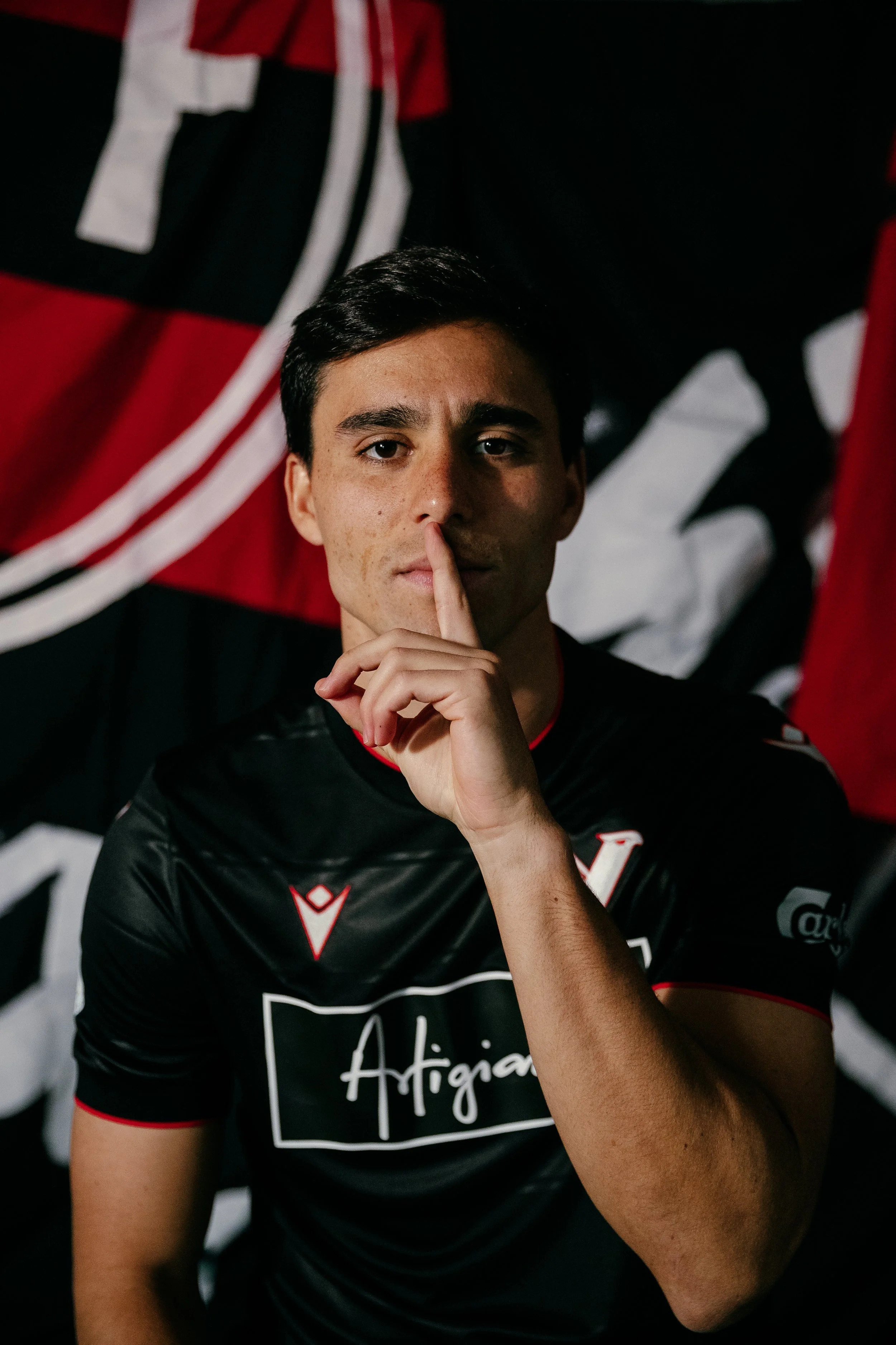 A young man with short dark hair and a serious expression, holding a finger to his lips in a shushing gesture, wearing a black sports jersey with a white and red logo and the word "Argentina" on it, standing in front of a black and red flag backgroun