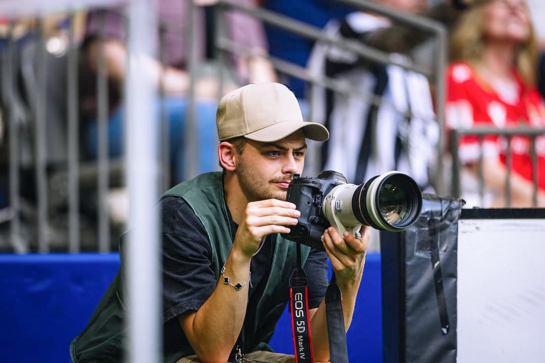 Young man with a camera taking photos at an outdoor event, seated near a railing with spectators in the background.