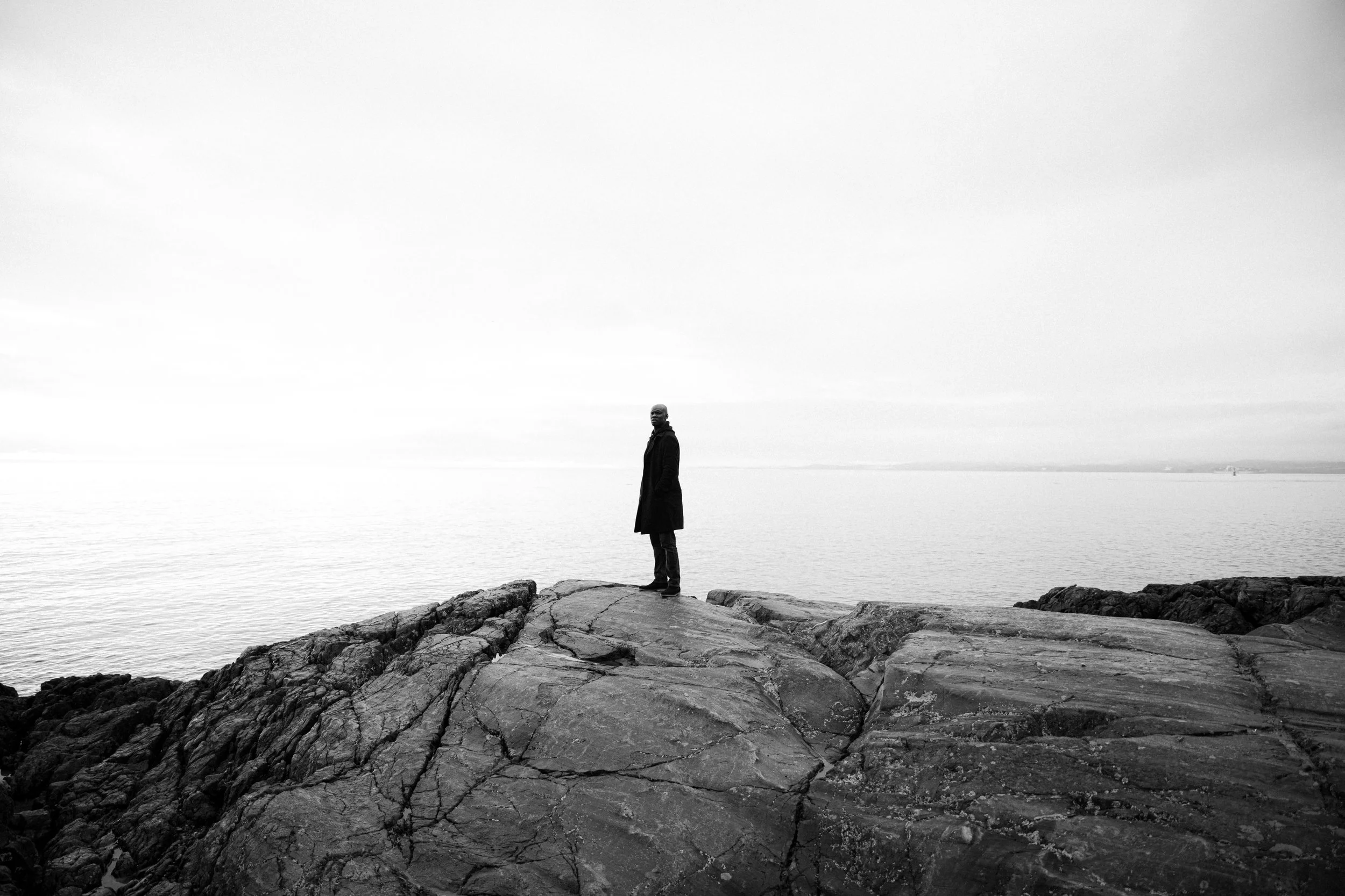 A person standing on rocks by the water with a cloudy sky in the background.