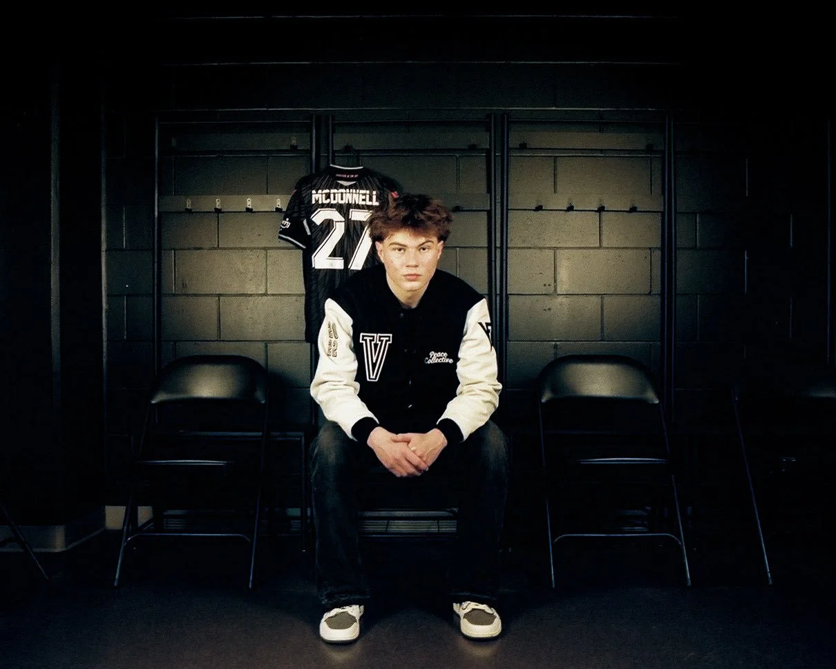 Young man sitting on a black chair in a locker room with a sports jersey hanging on the wall behind him.