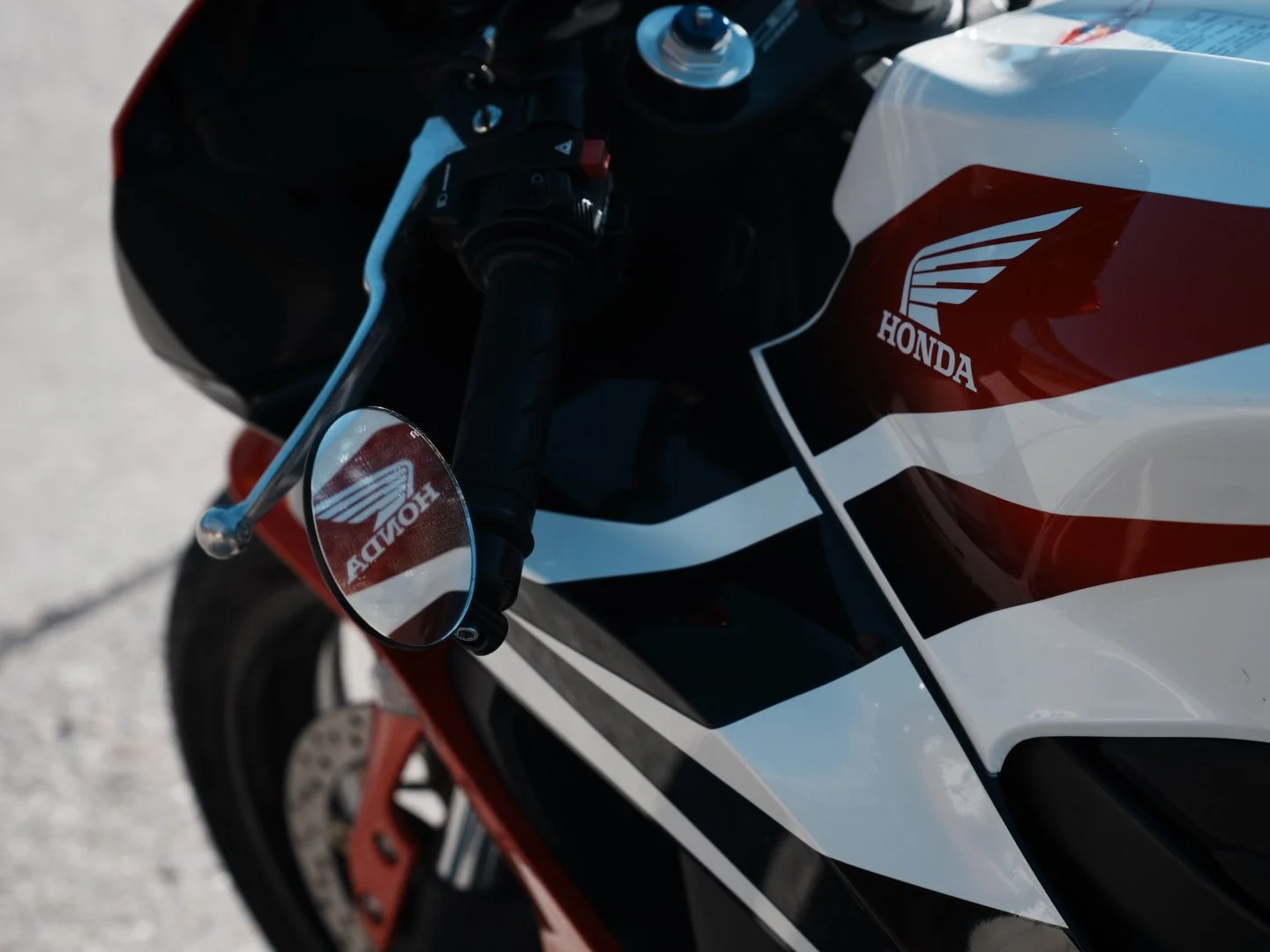 Close-up of a Honda motorcycle with a red and white color scheme, showing handlebars, mirror, and the Honda logo on the fairing.