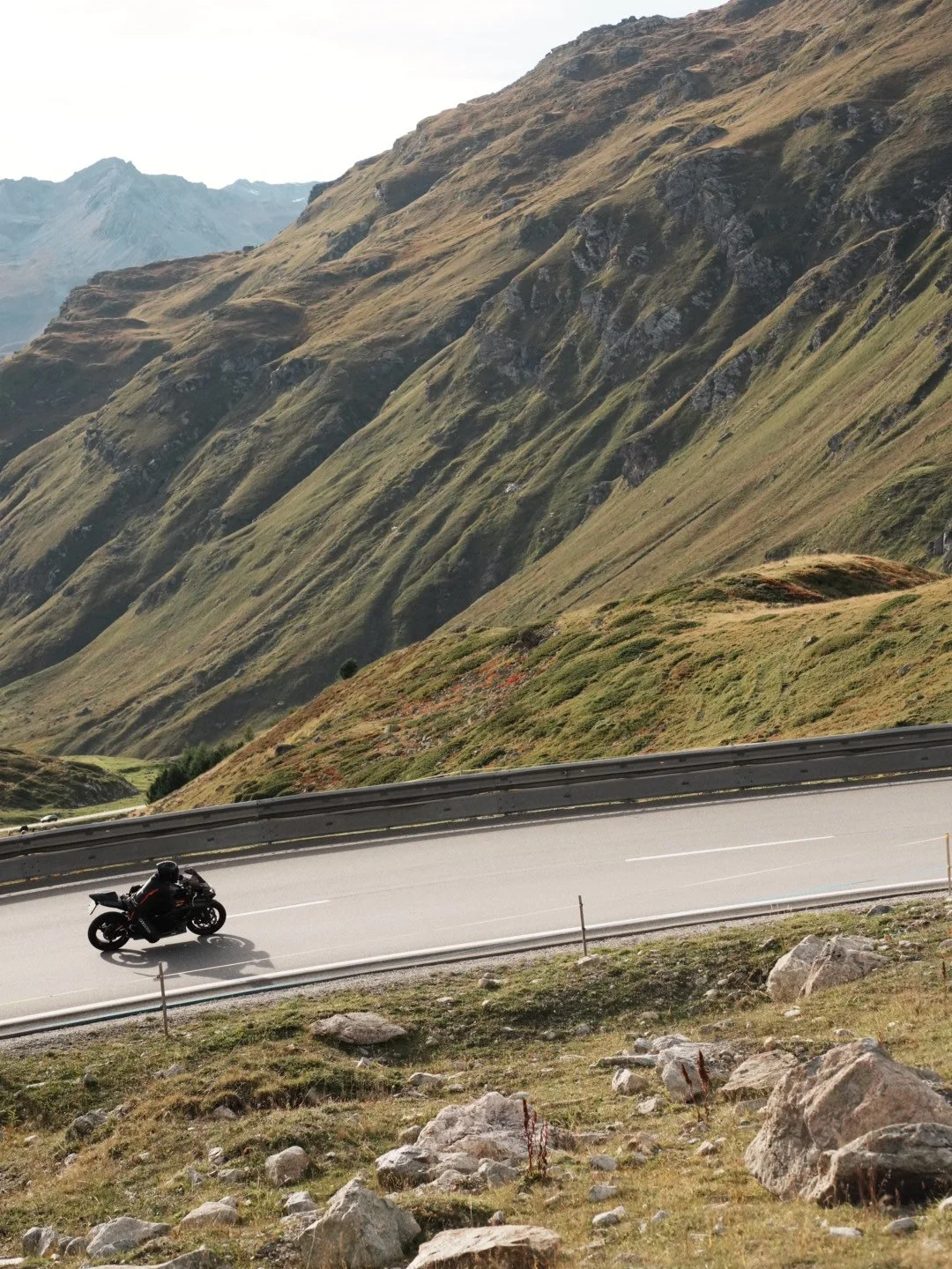A black motorcycle riding on a winding mountain road surrounded by steep grassy slopes and rocky terrain.