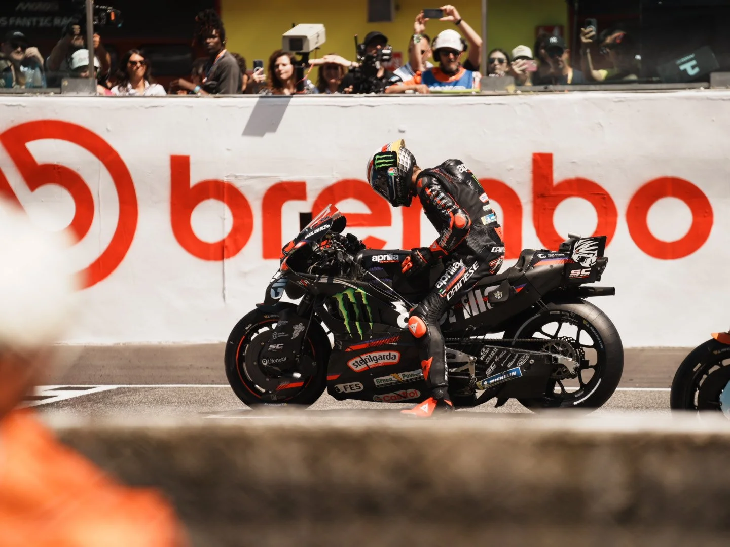A professional motorcycle racer in full gear, including a helmet and racing suit, standing next to a black and green motorcycle on a racetrack, with spectators watching behind a barrier and a large billboard with red and white text in the background.
