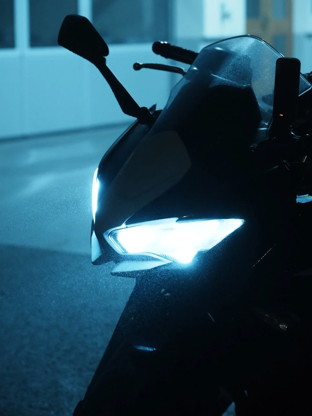 Close-up of the front of a black motorcycle with bright headlights turned on, parked indoors in low light.