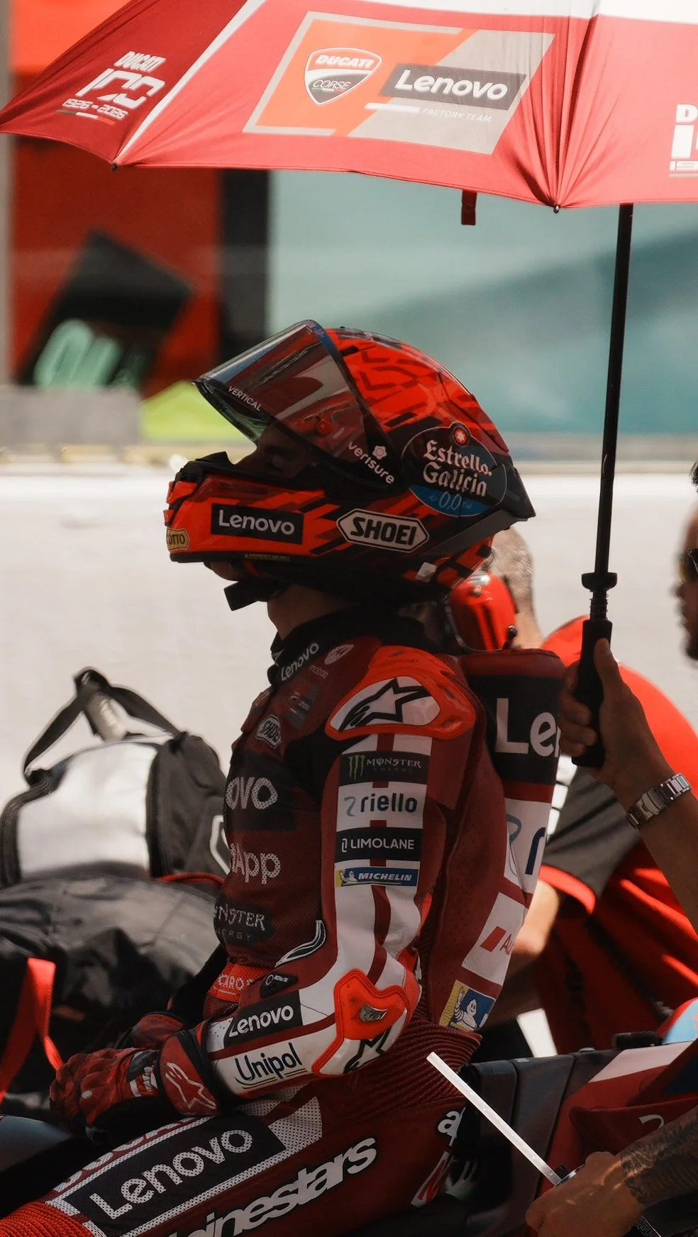 Motorsport athlete wearing a red racing suit and helmet, seated on a motorcycle, holding an umbrella for shade.