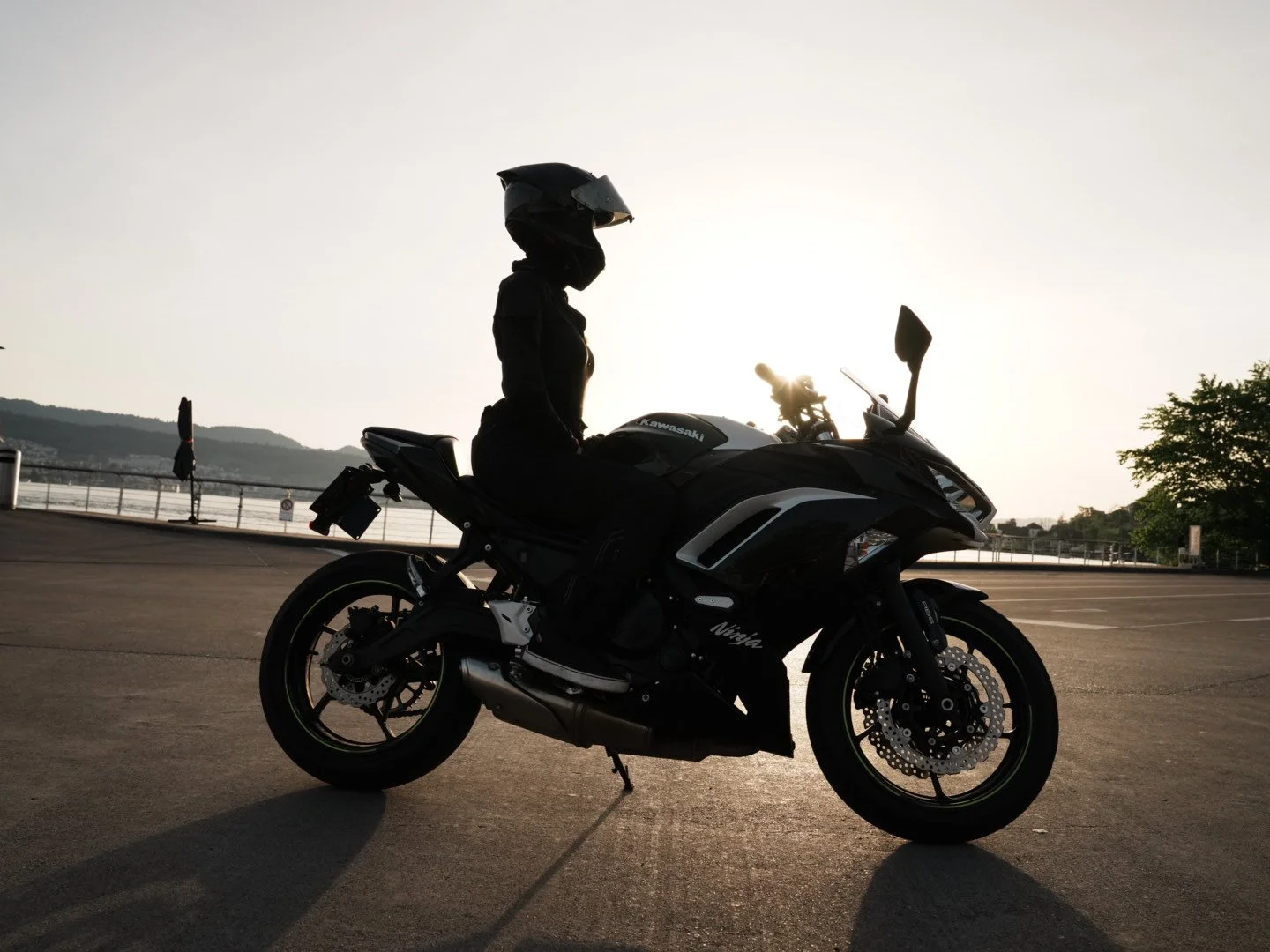 Silhouette of a motorcycle rider wearing a helmet, sitting on a black Kawasaki Ninja motorcycle, with a sunset and hills in the background.