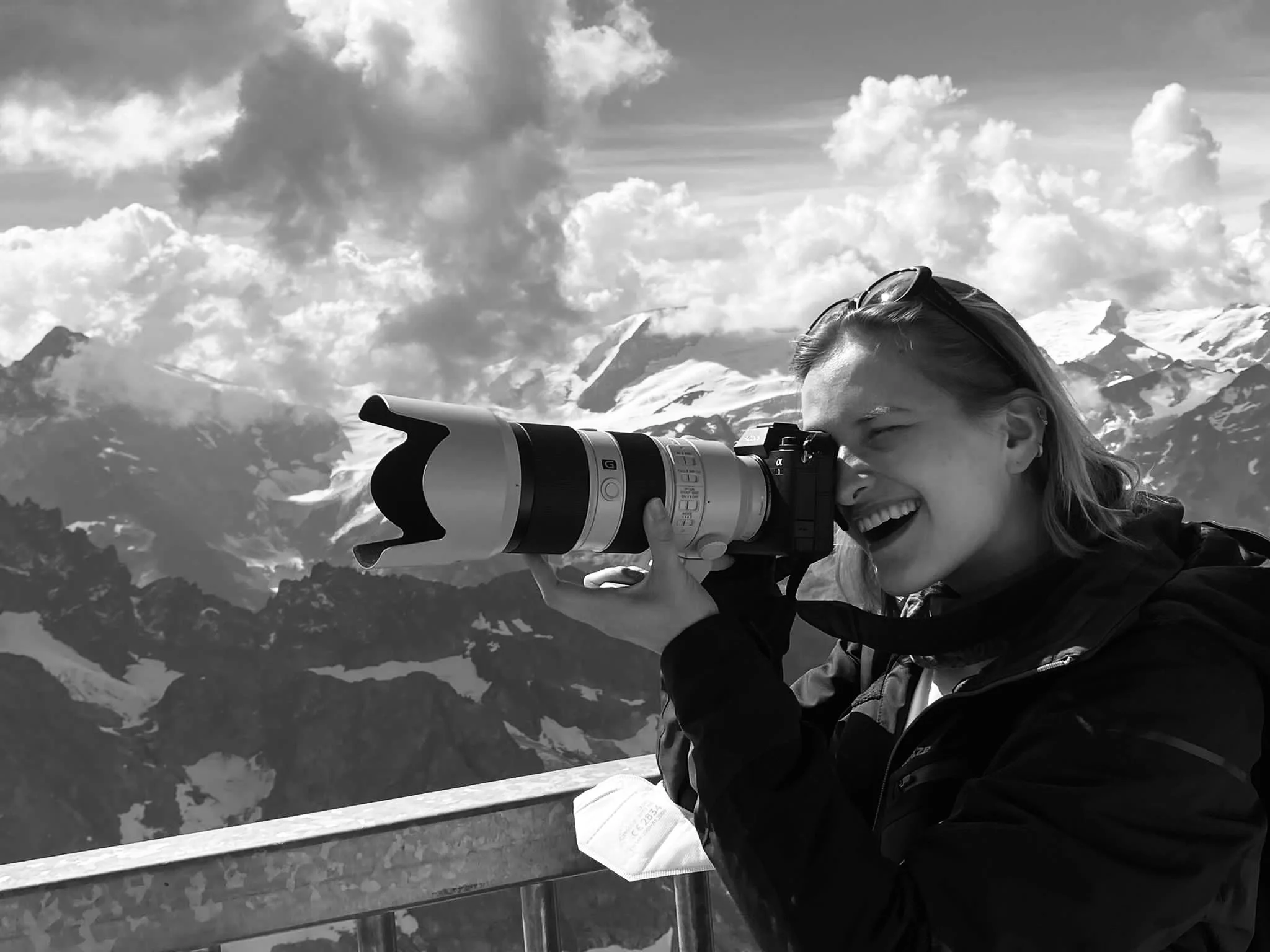 A woman with sunglasses on her head is seen using a large camera with a telephoto lens to take a photograph. She is outdoors in a mountainous area with snow-capped peaks and clouds in the sky.